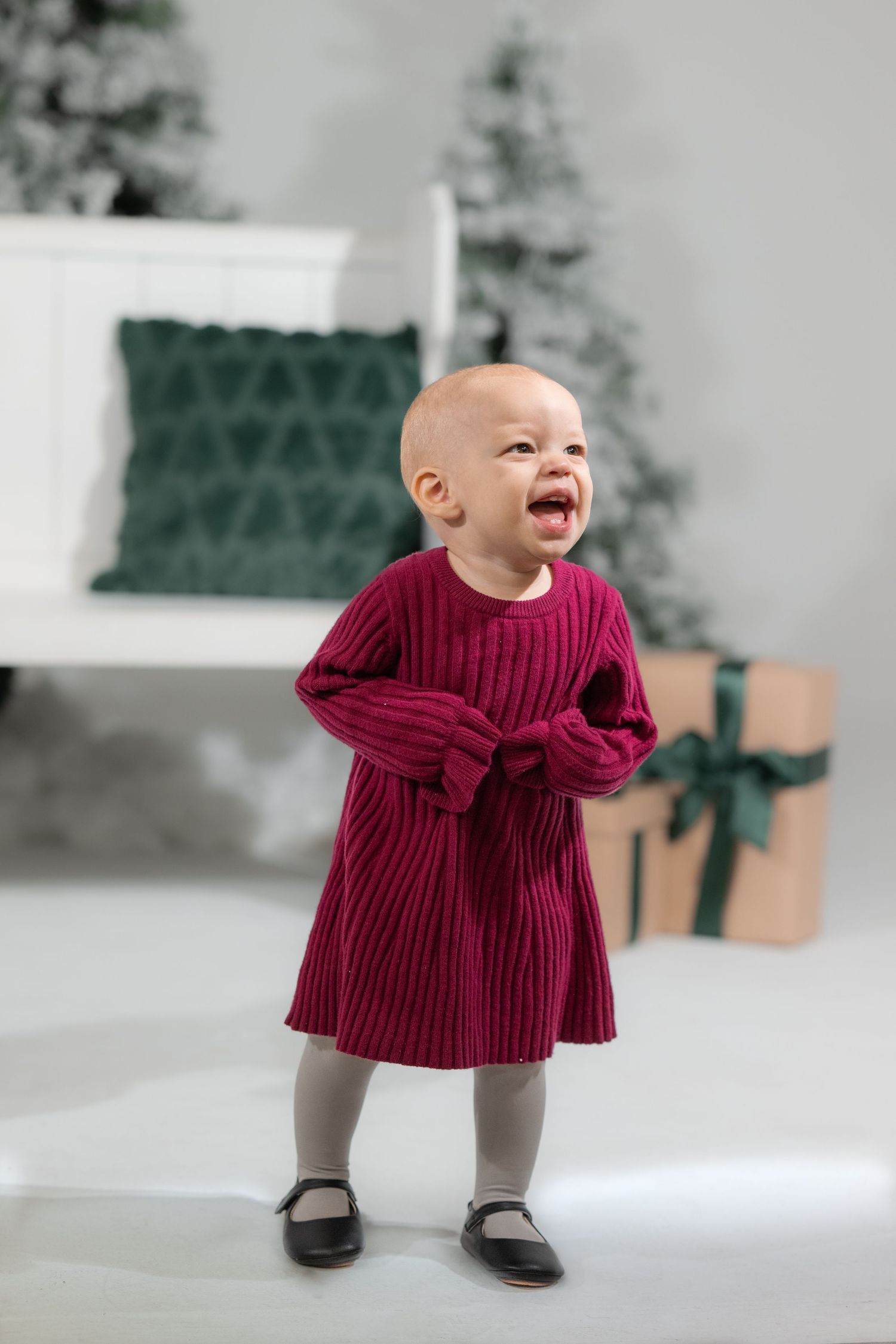 Baby in burgundy dress crawls and plays on white studio floor.