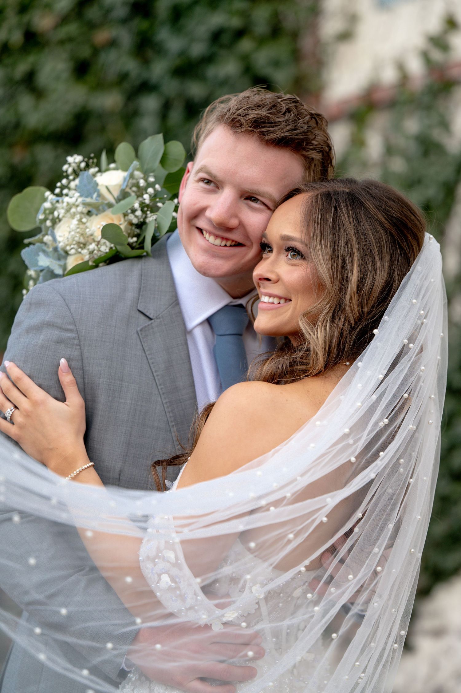 Smiling bride and groom embrace outdoors, bride wearing veil and holding white floral bouquet, groom in gray suit.