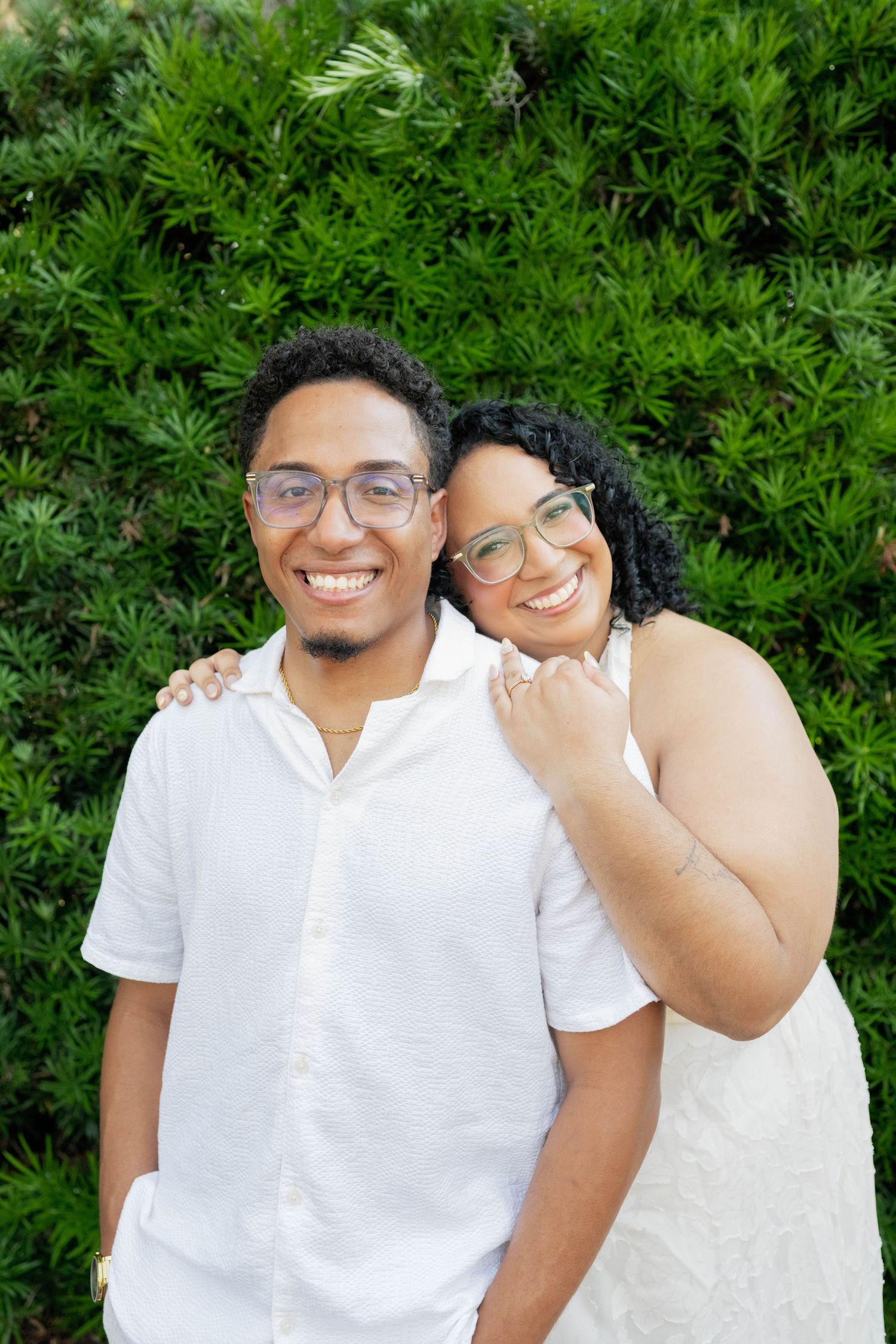 Smiling couple in matching white outfits and glasses posing together in front of lush green shrubs outdoors.