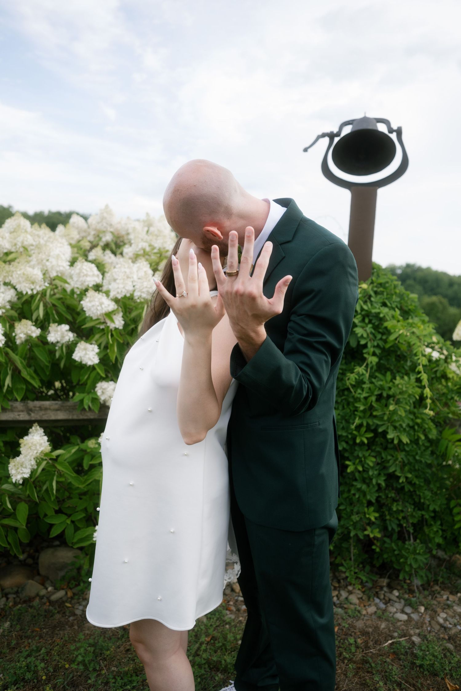 Newlywed couple kissing while showing off wedding rings, wearing white dress and green suit near white hydrangeas.