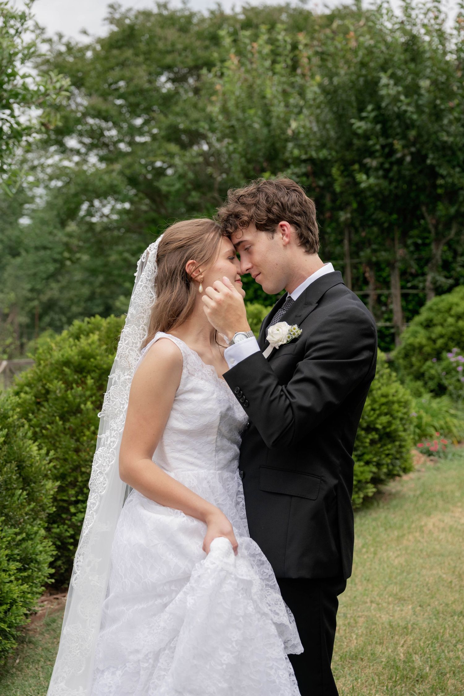A bride and groom share an intimate forehead touch moment in a lush green garden, wearing classic white gown and black tuxedo.