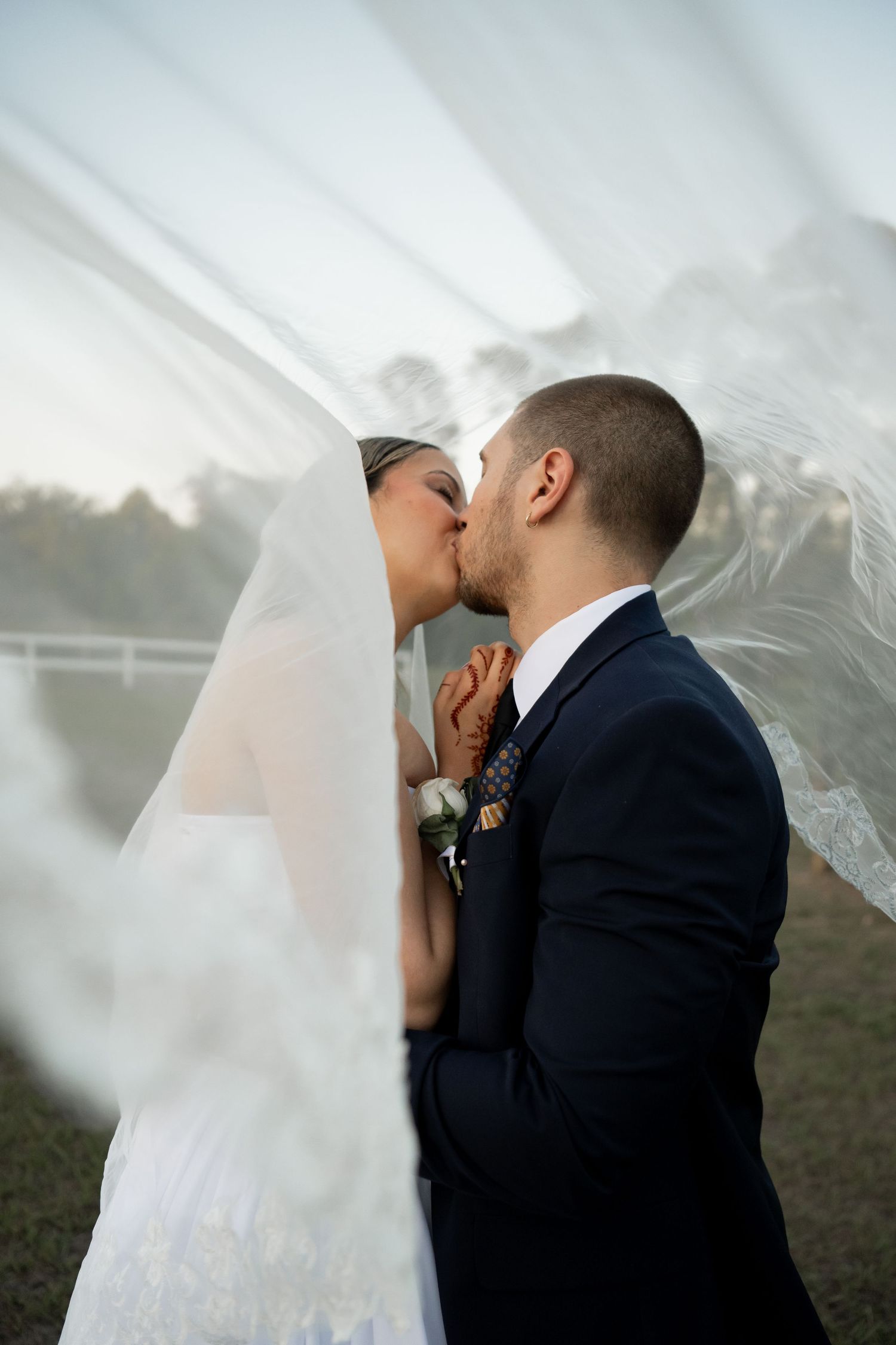 Artistic wedding portraits of couple kissing beneath flowing veil at sunset.