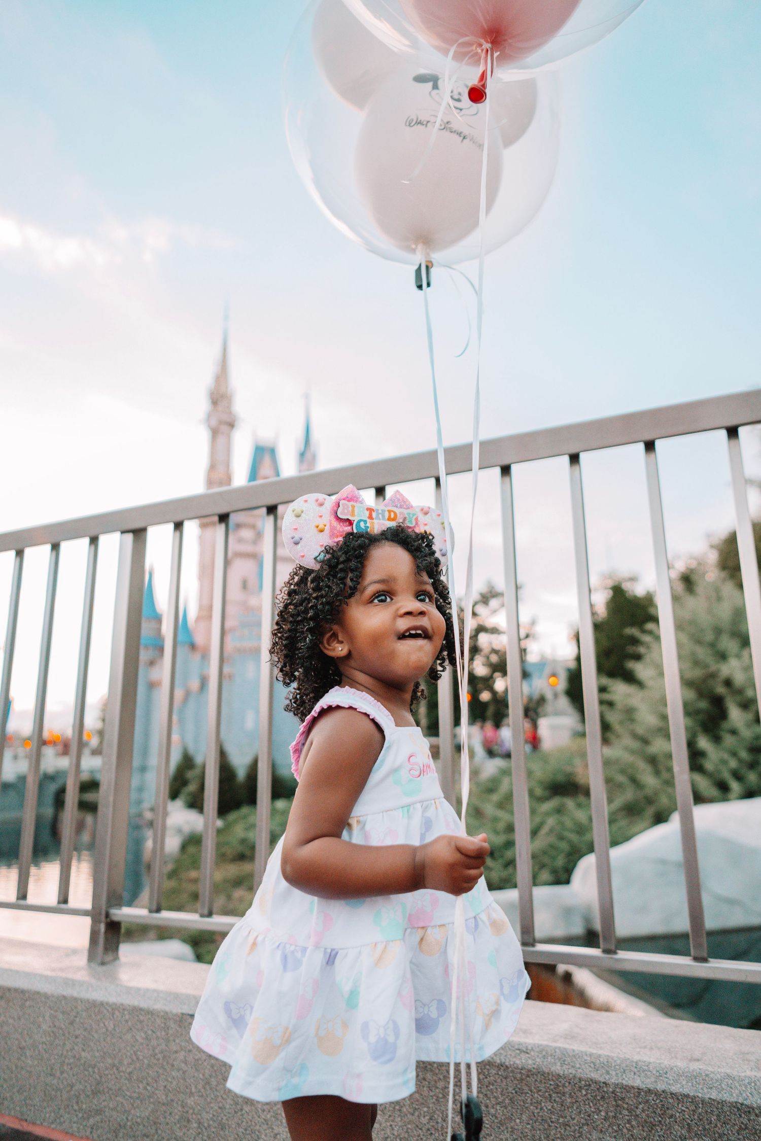 Young child in white dress holding pink balloons at Disney World with castle in background.