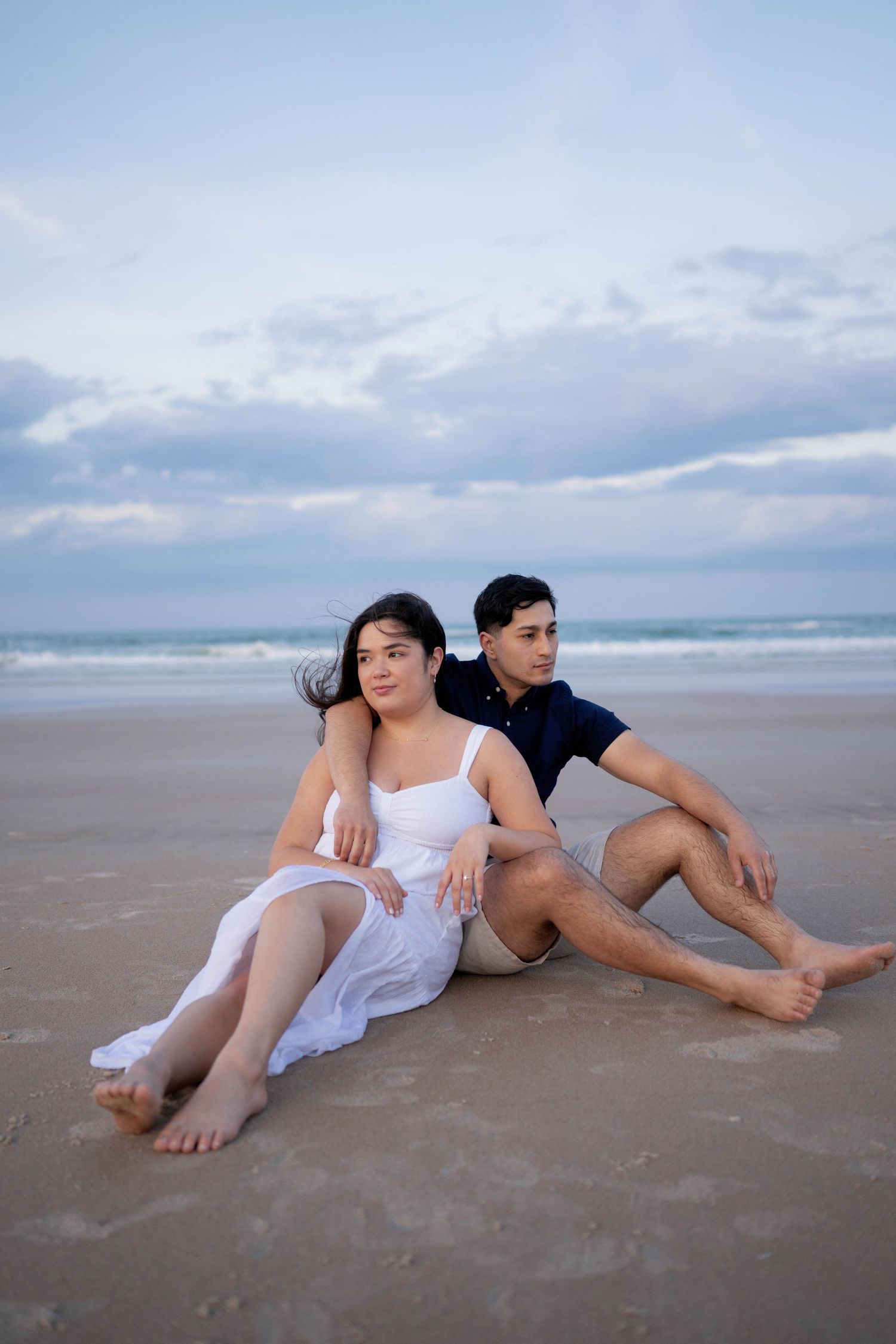 A couple sits together on a sandy beach at dusk, woman in white dress leaning against man in navy shirt.