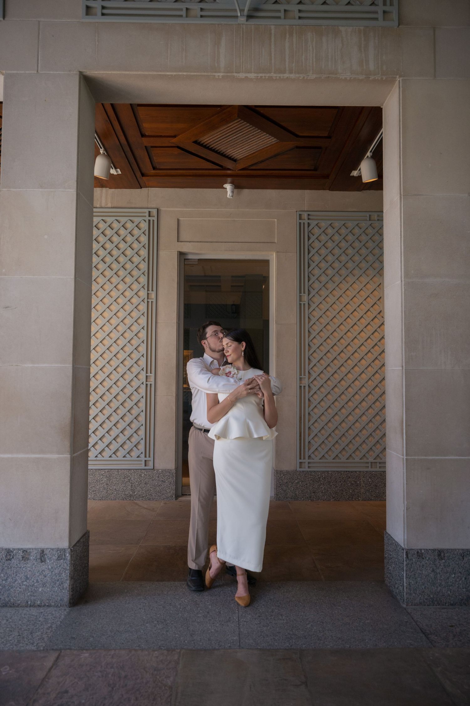 Couple embracing in a stone archway with a wood coffered ceiling, she in a white peplum top and skirt, he in a white shirt and khakis.