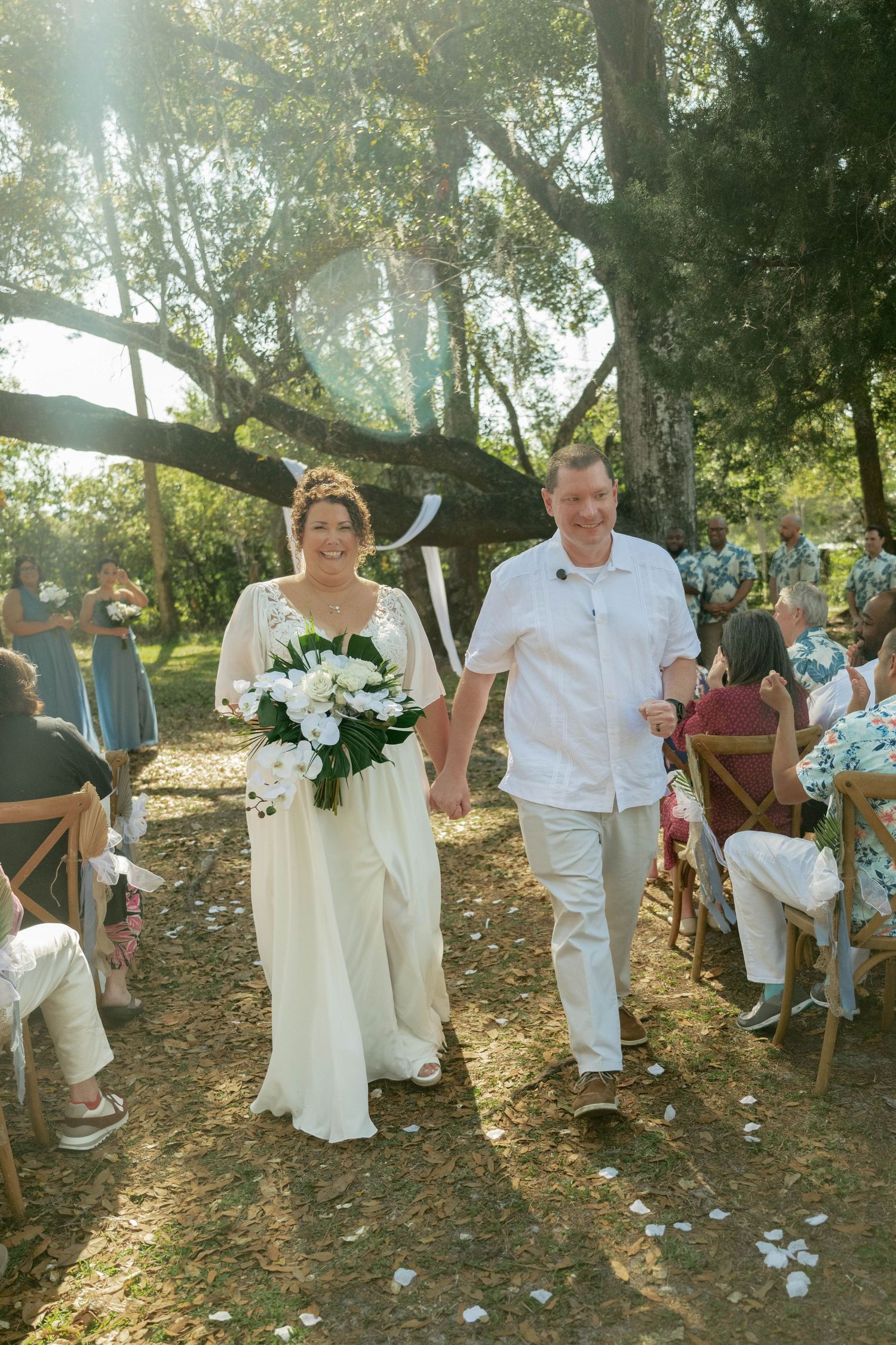 Bride and groom walk hand-in-hand down outdoor aisle after wedding ceremony under large oak trees.