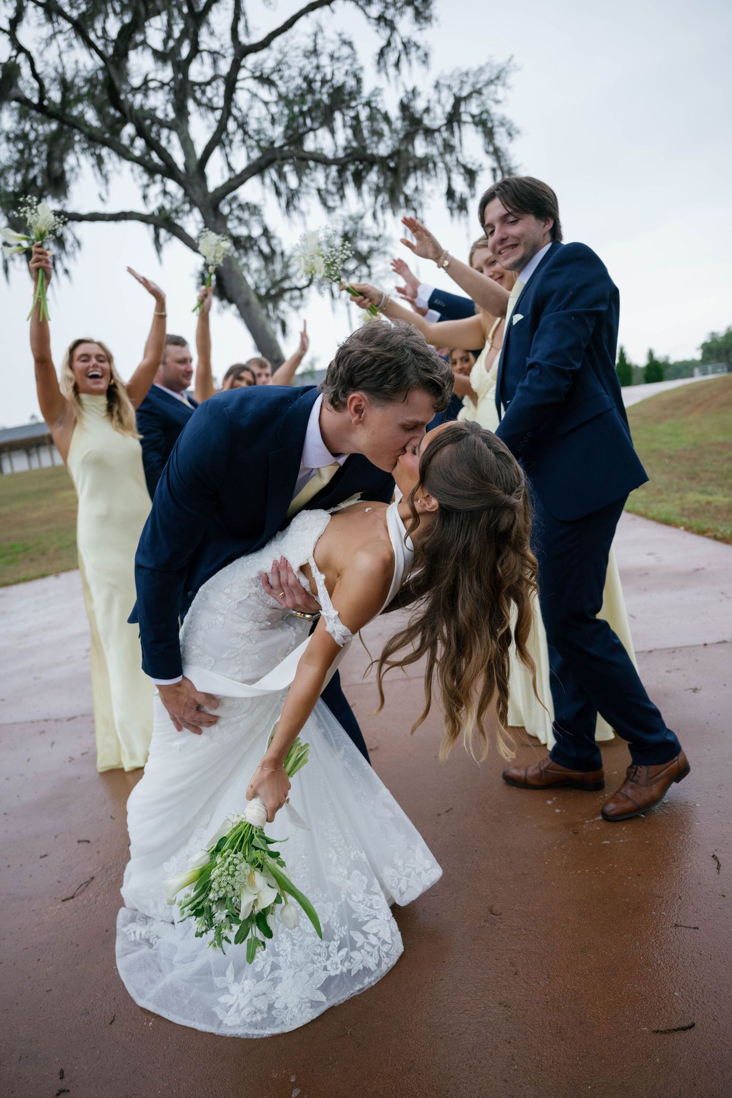 Groom dips bride for a kiss while wedding party celebrates with raised arms outdoors.