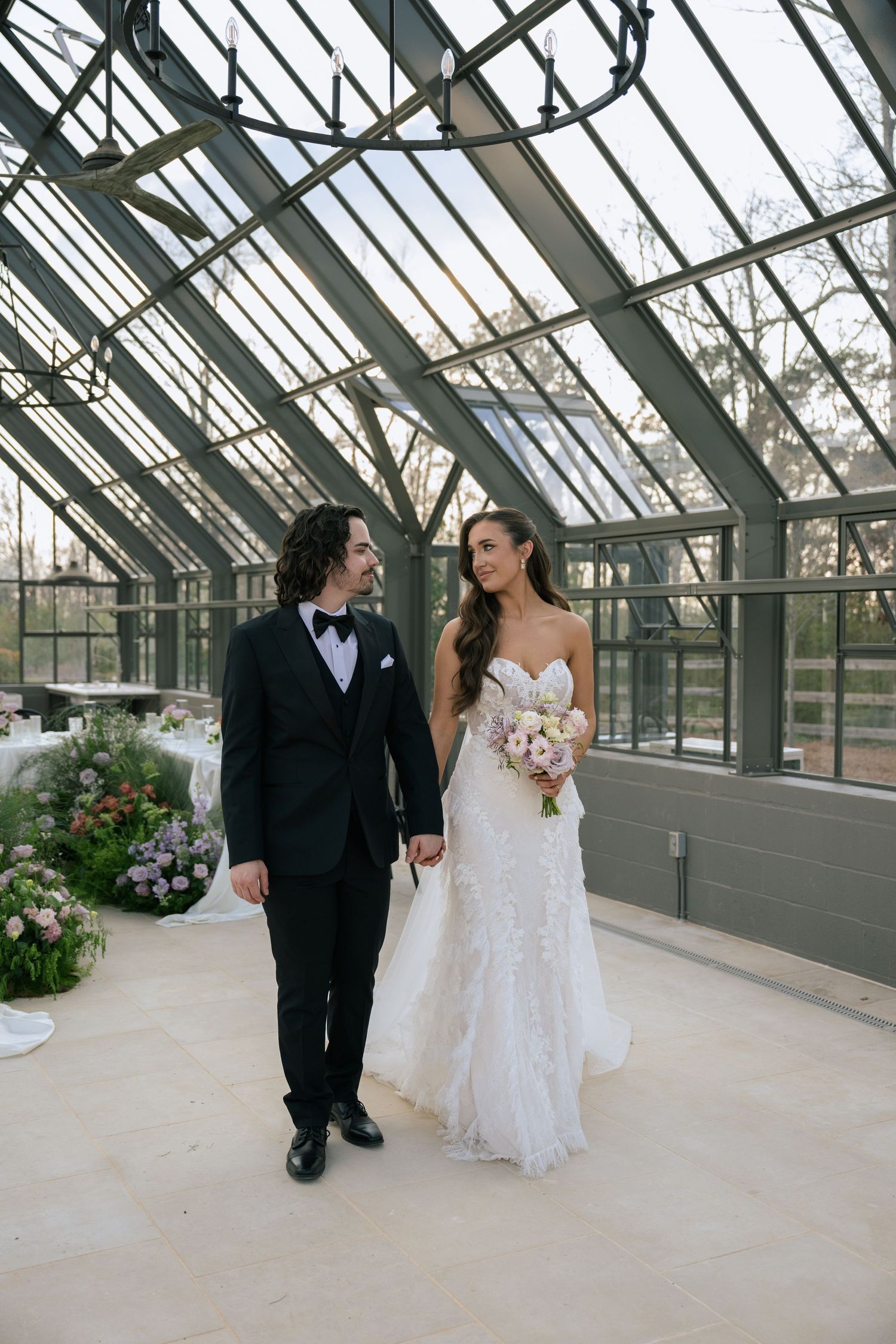 Bride and groom holding hands in a stunning glass greenhouse wedding venue, bride holding a pastel bouquet.