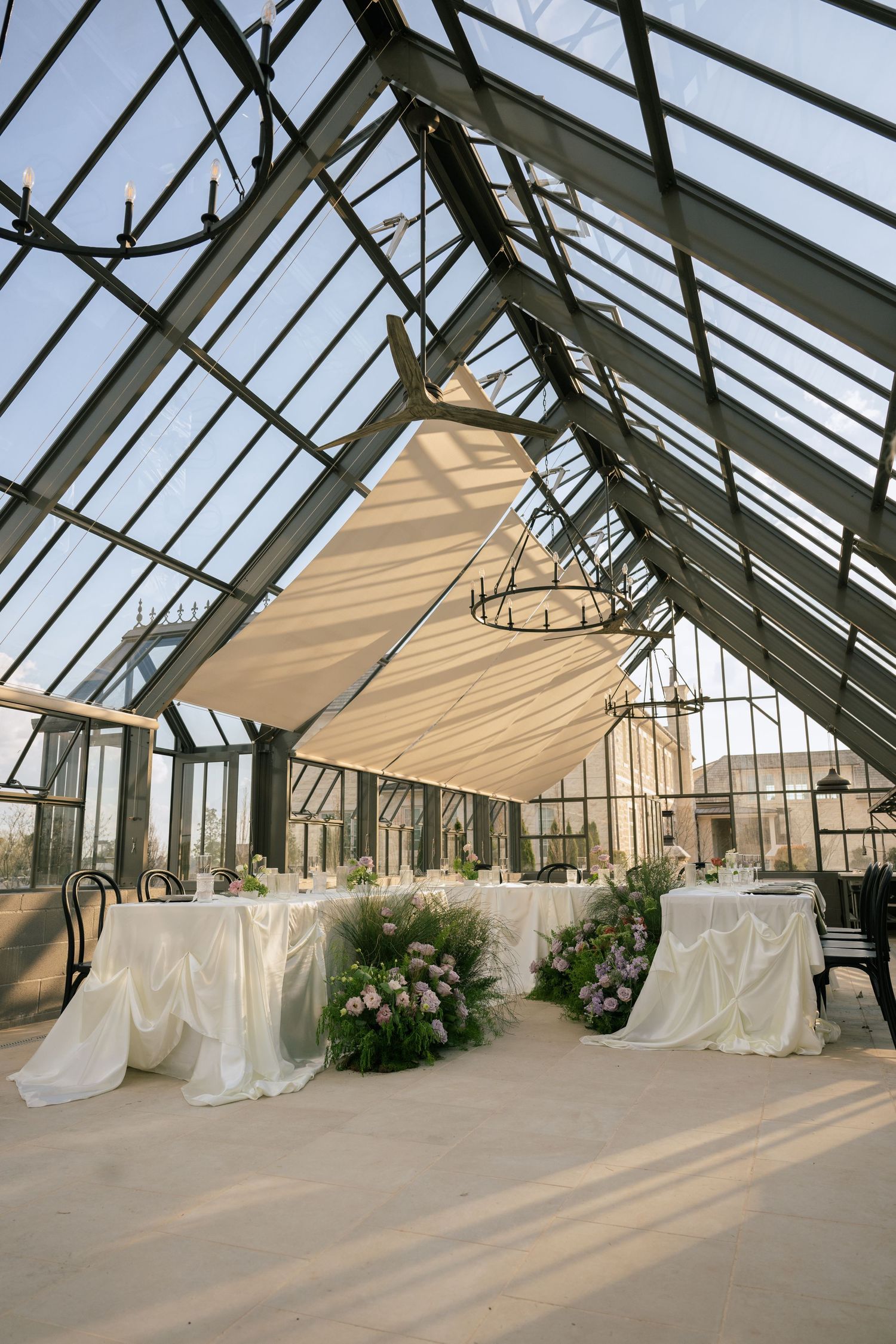 Elegant wedding reception setup in a glass greenhouse venue with white draped tables and floral arrangements.