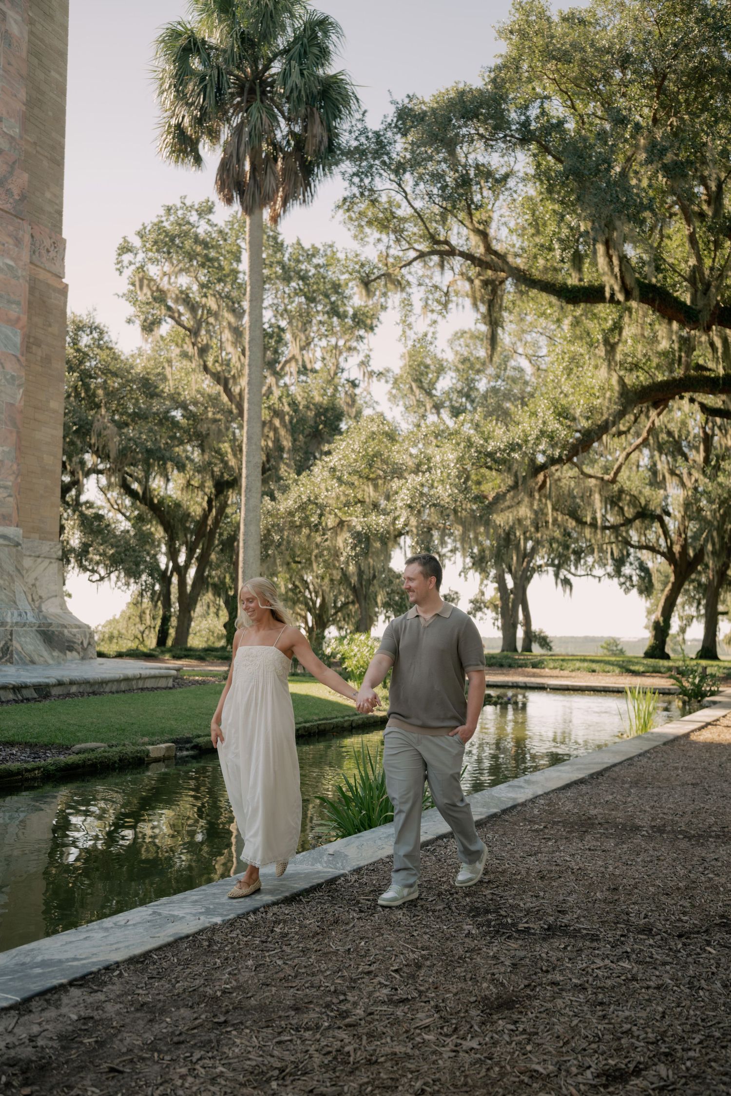Couple holding hands walking beside a reflective pond surrounded by moss-draped oak trees and palm trees.
