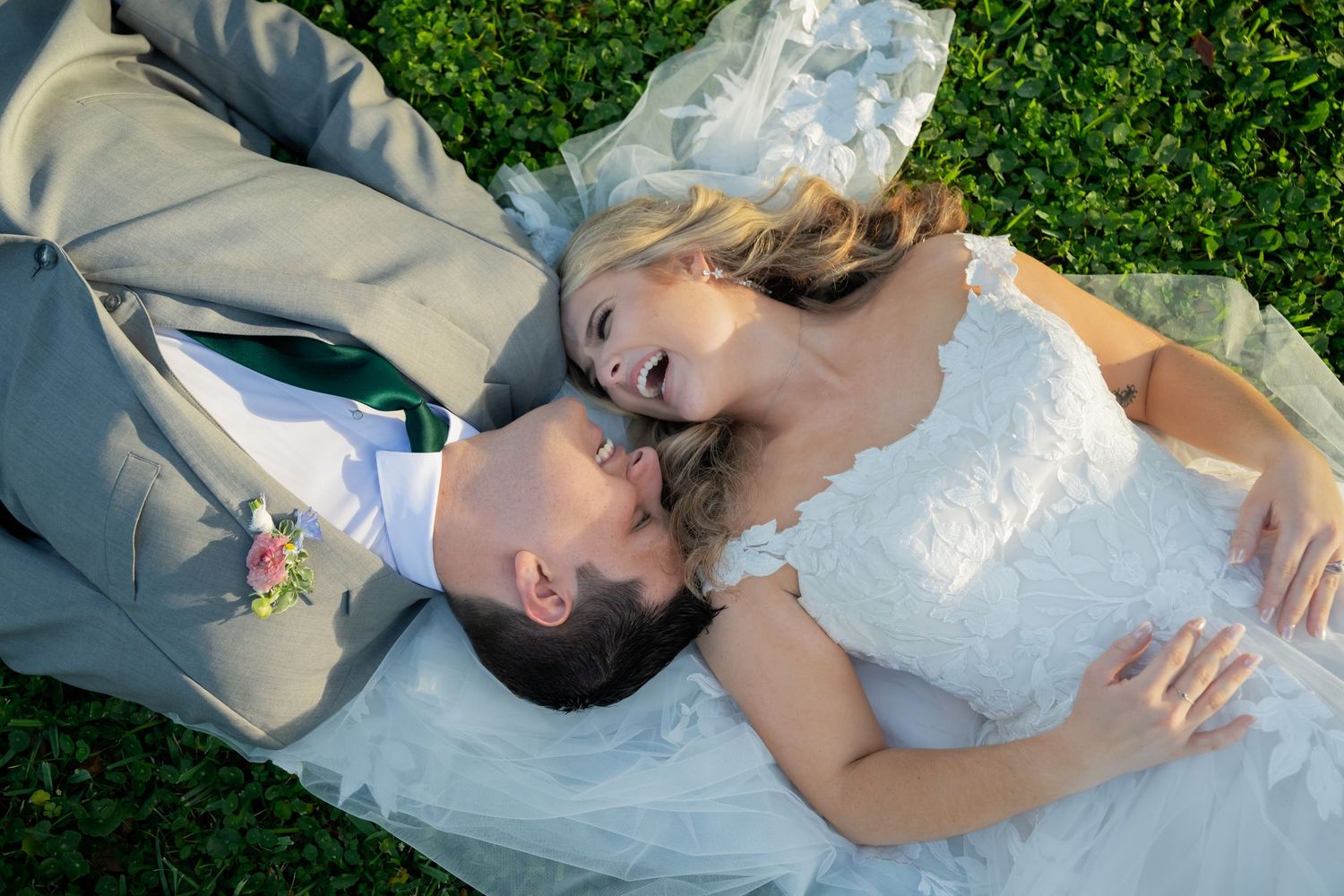 Romantic couple in wedding attire lying together on green grass during outdoor photo session.
