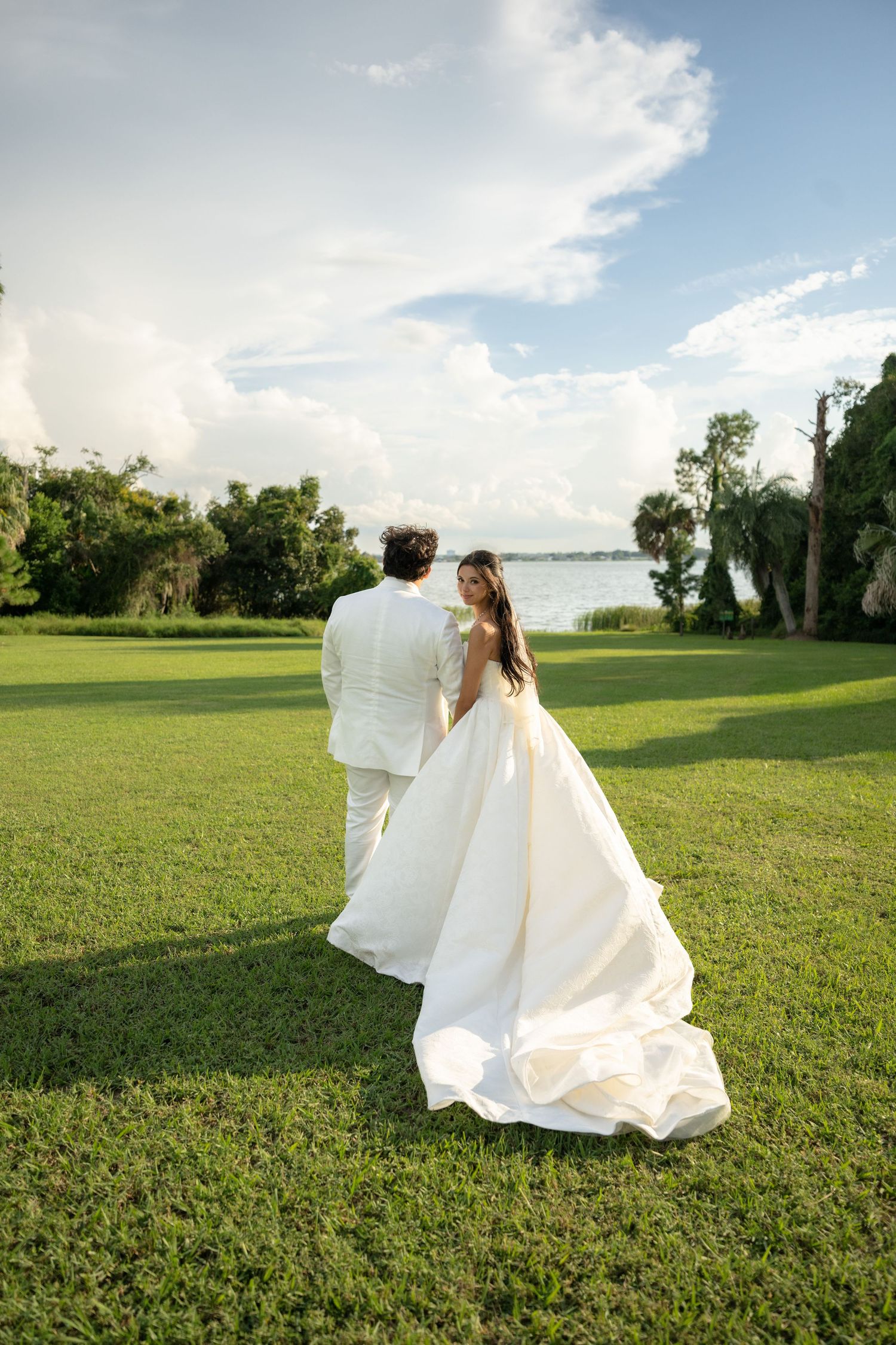 Bride in a flowing white gown and groom in a cream suit walk across a sunny green lawn toward a lakeside under a bright blue sky.