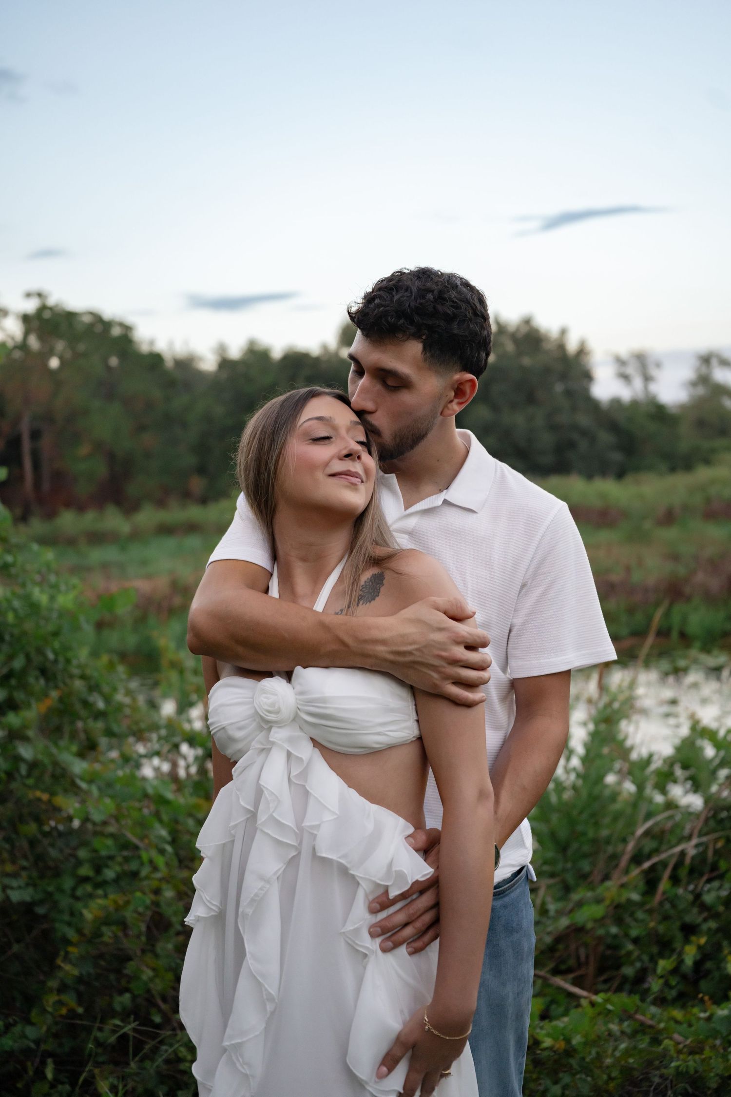 Romantic couple embracing outdoors, man kissing woman's temple, both in white attire with lush greenery background.