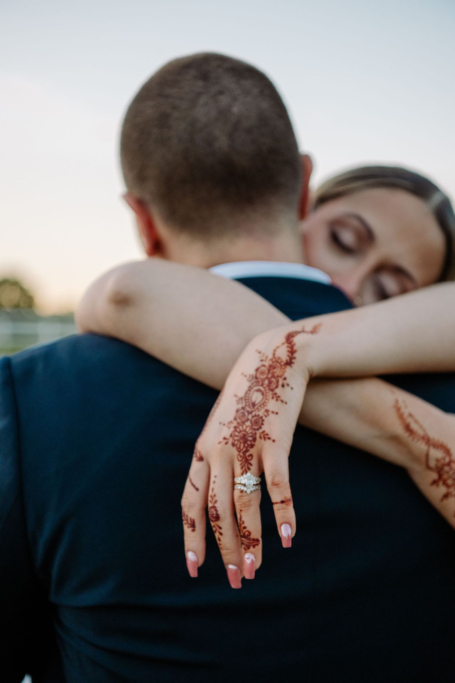 Close-up of henna-decorated hand with engagement ring resting on dark formal wear.