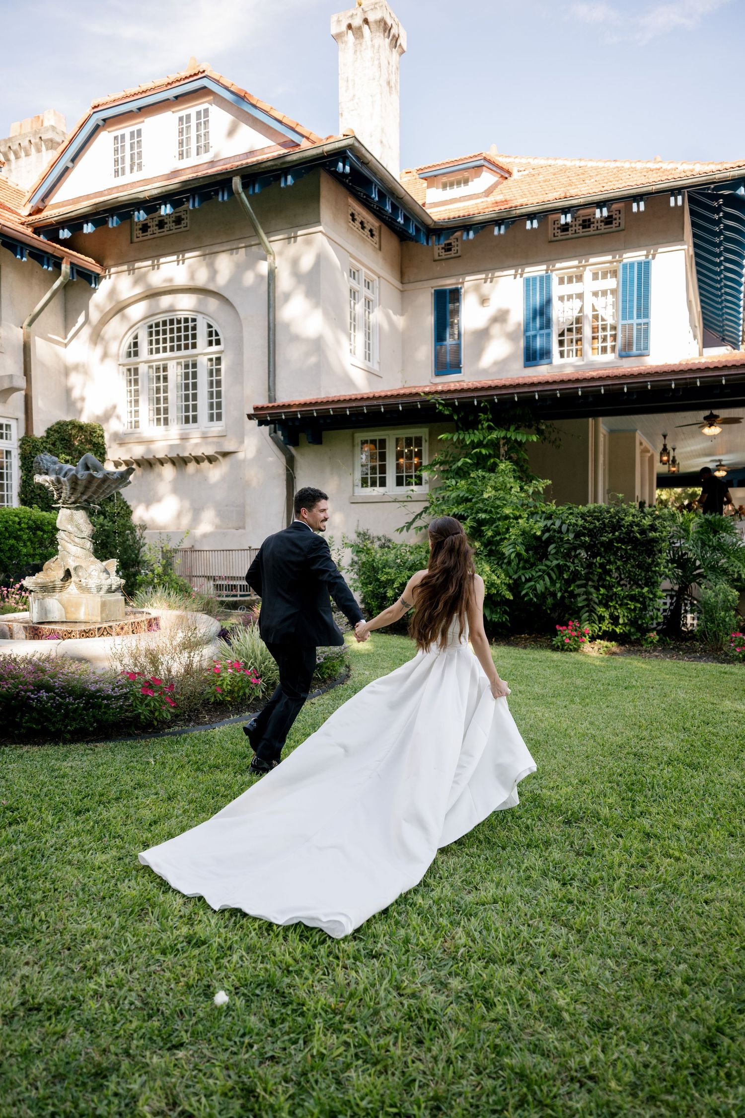 Bride in a flowing white gown with a long train holds hands with the groom while walking across a green lawn toward a historic estate.