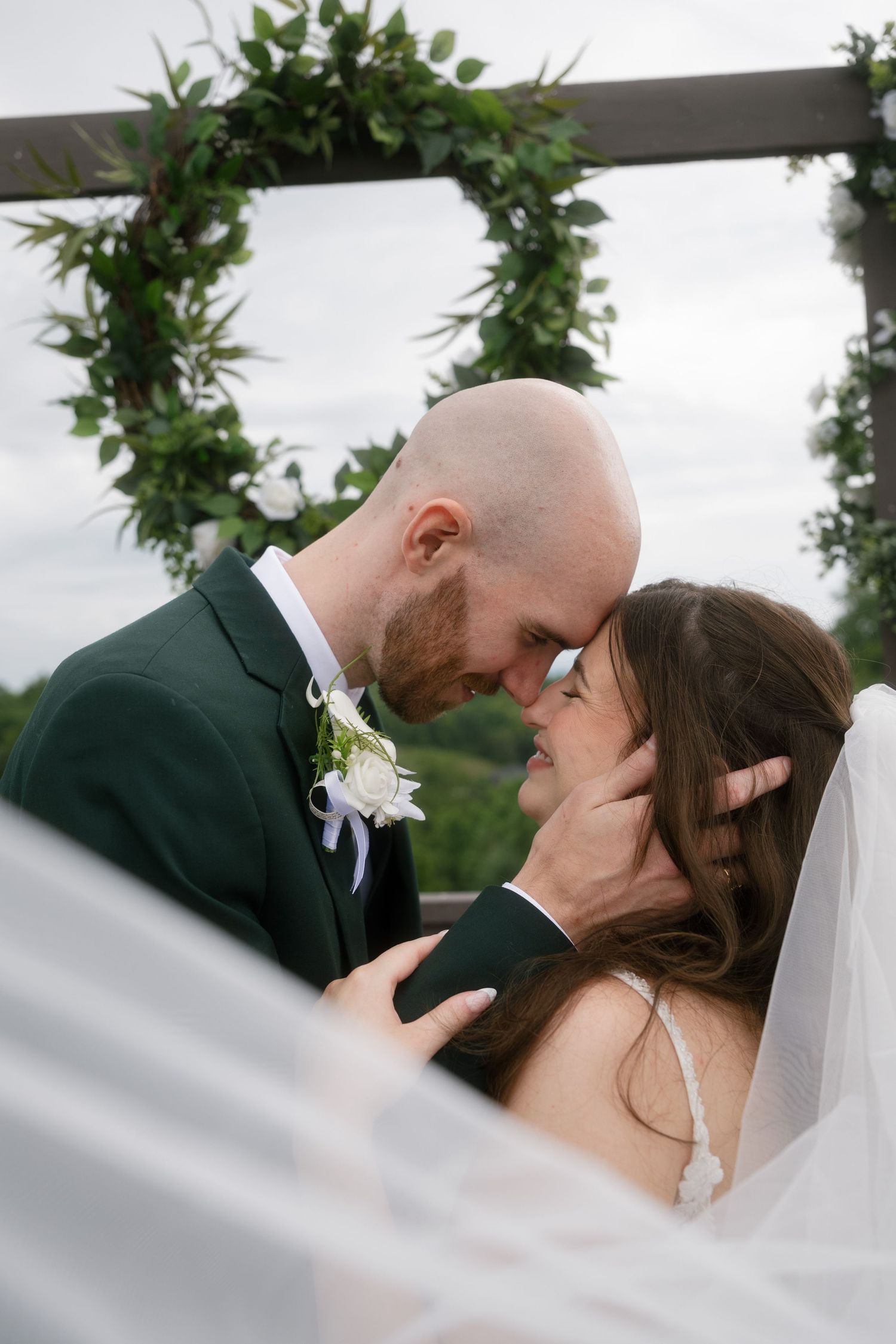 Bride and groom touching foreheads tenderly at outdoor wedding ceremony with greenery wreath arch backdrop.