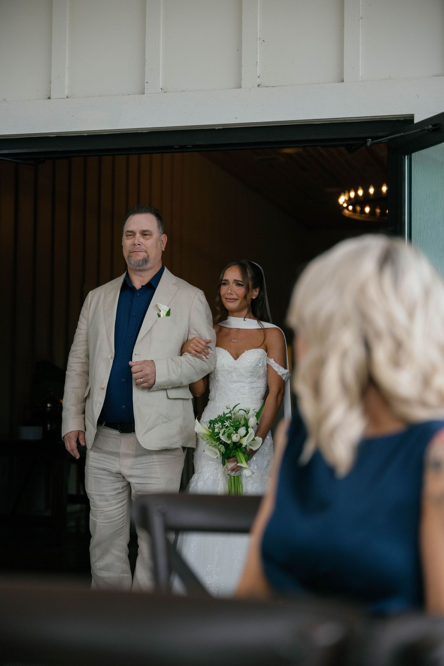 Bride in white gown holding bouquet walks arm-in-arm with escort through venue doors at elegant wedding ceremony.
