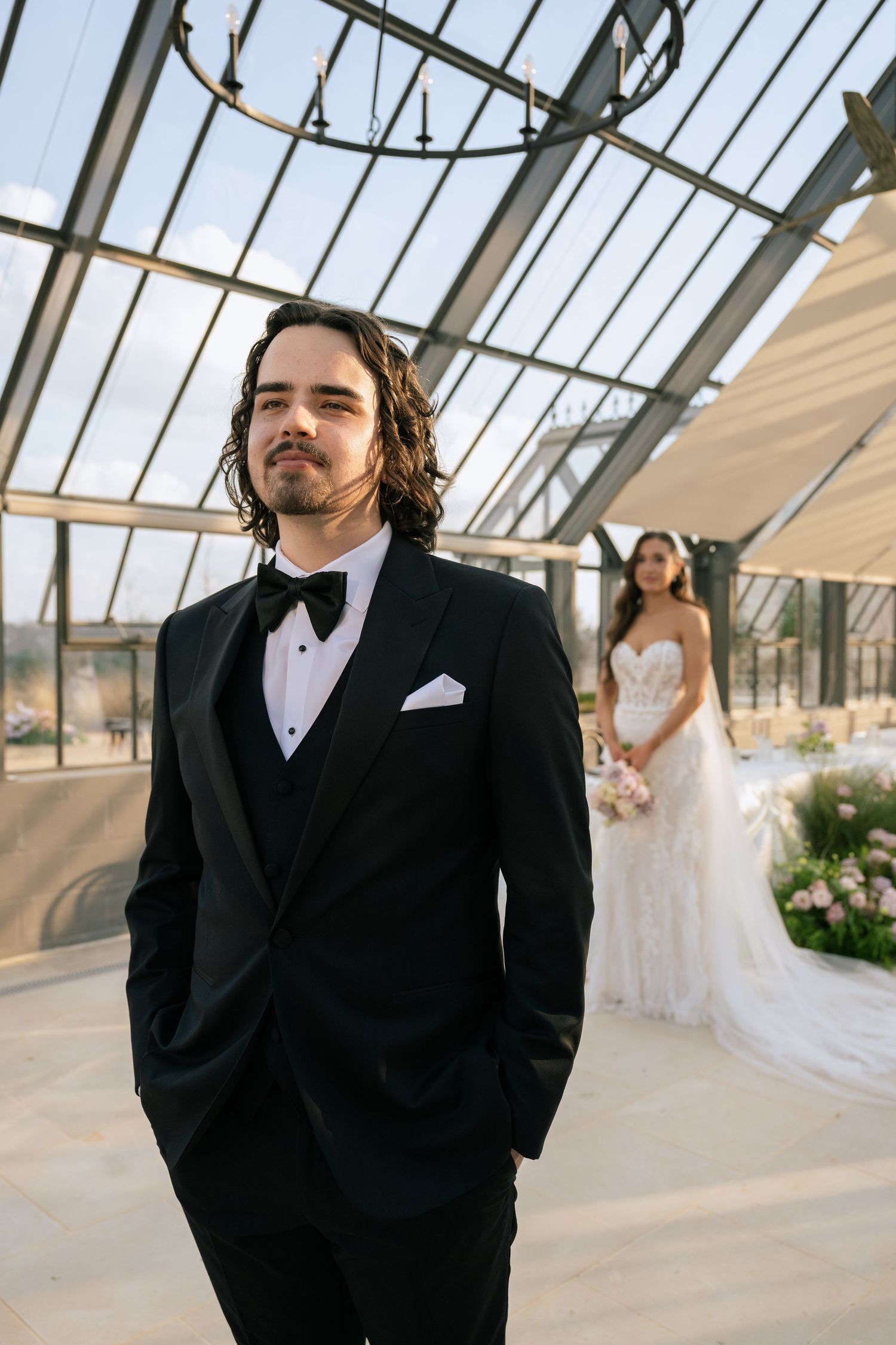 Groom in black tuxedo stands in foreground while bride in white lace gown holds bouquet in glass venue.