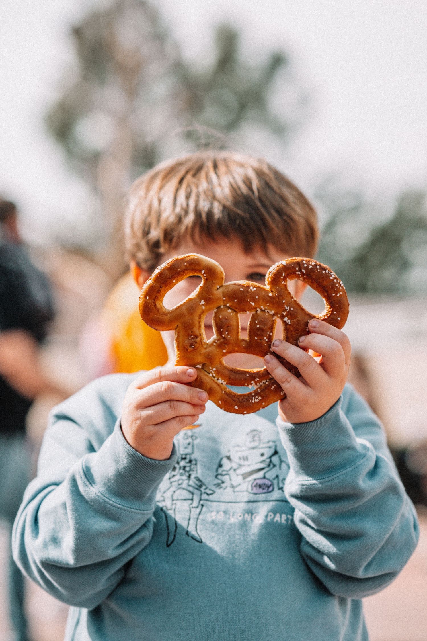 Child holding up Mickey Mouse shaped pretzel against blurred background.