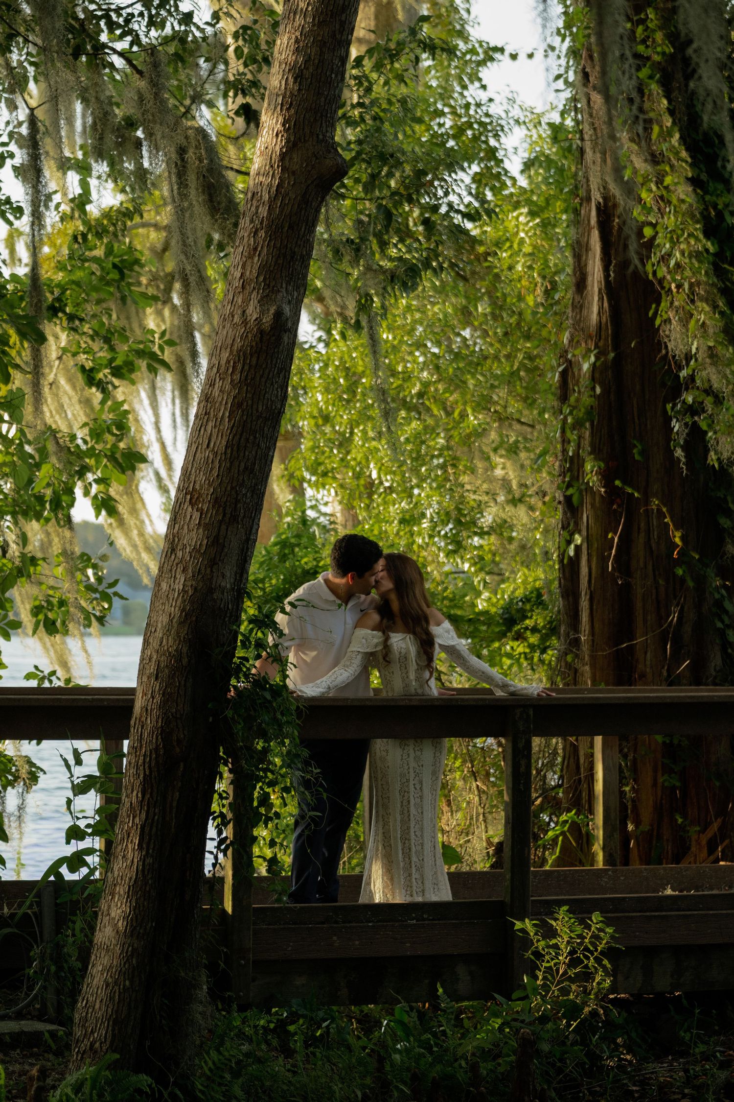 Couple sharing a romantic kiss on a wooden deck surrounded by lush green trees with Spanish moss near a lake.