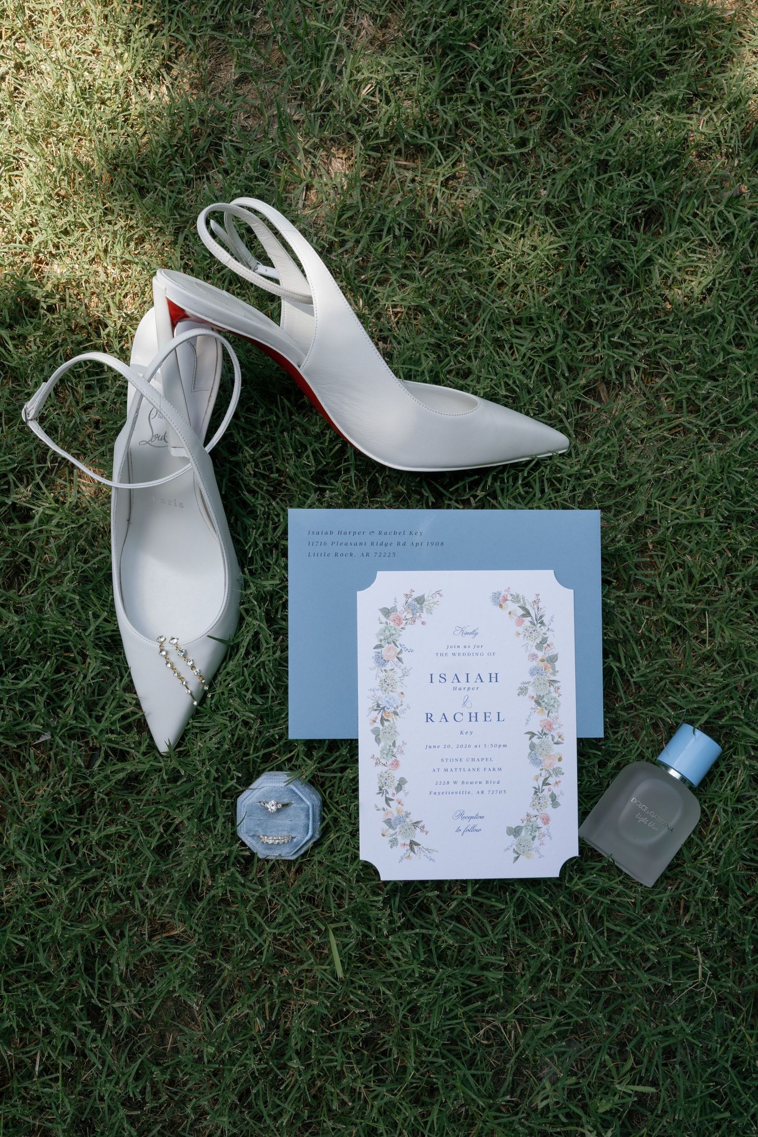 White Christian Louboutin heels, floral wedding invitation for Isaiah and Rachel, diamond ring, and blue perfume on grass.