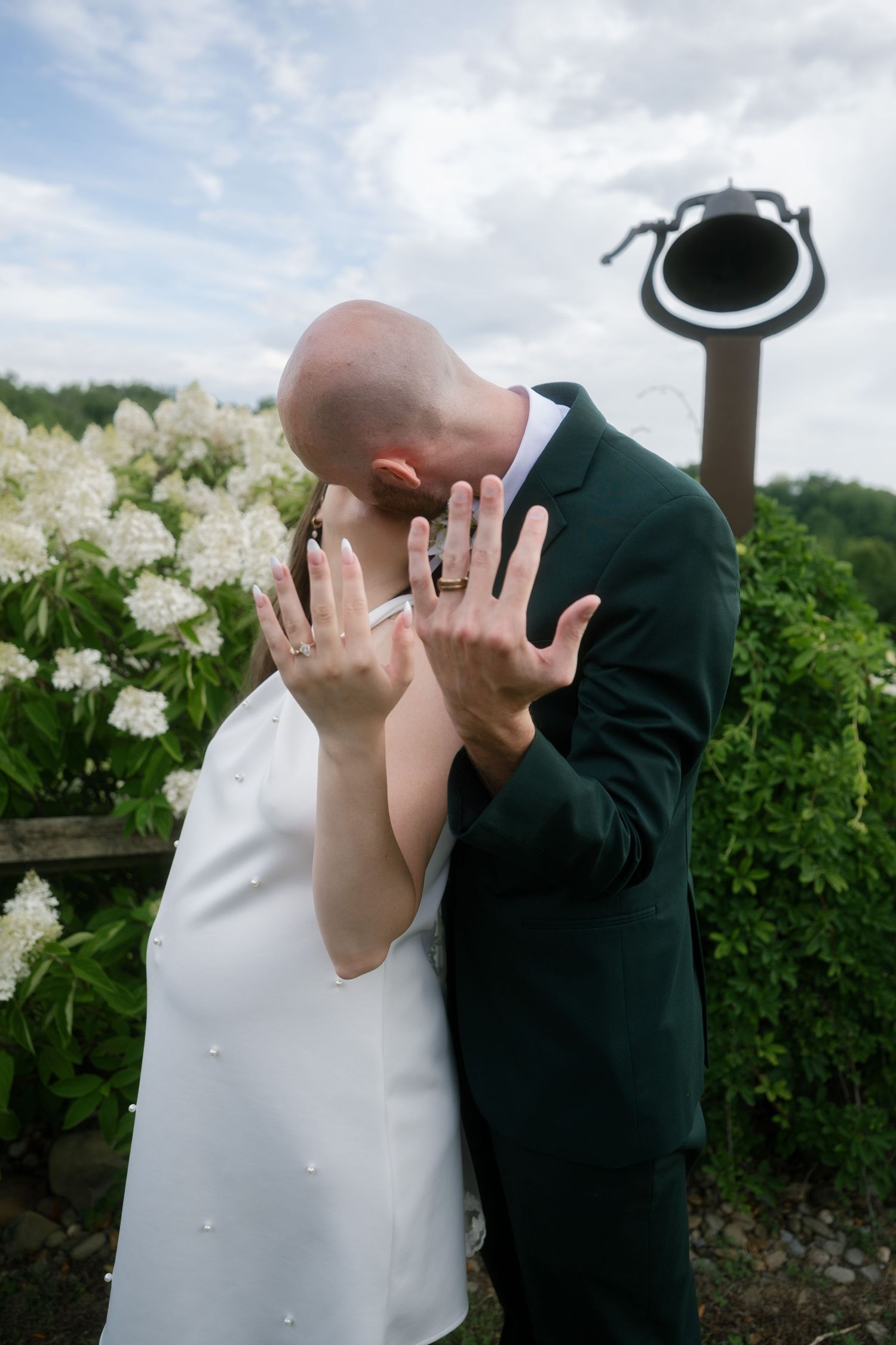 Newlywed couple kissing while showing off wedding rings, wearing white dress and green suit, with white flowers outdoors.