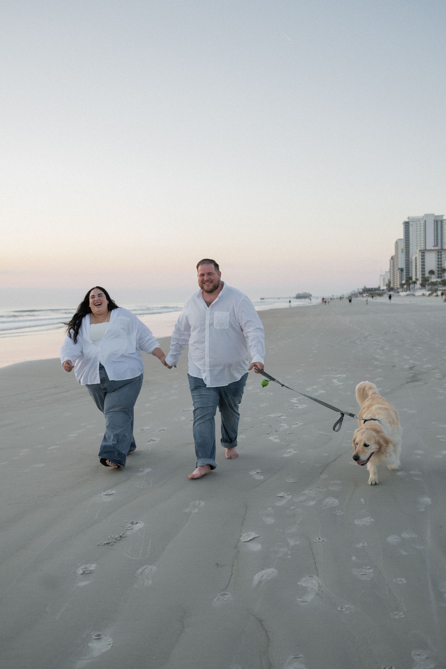 Couple in white shirts walking a golden retriever on a sandy beach at dusk with city skyline in background.