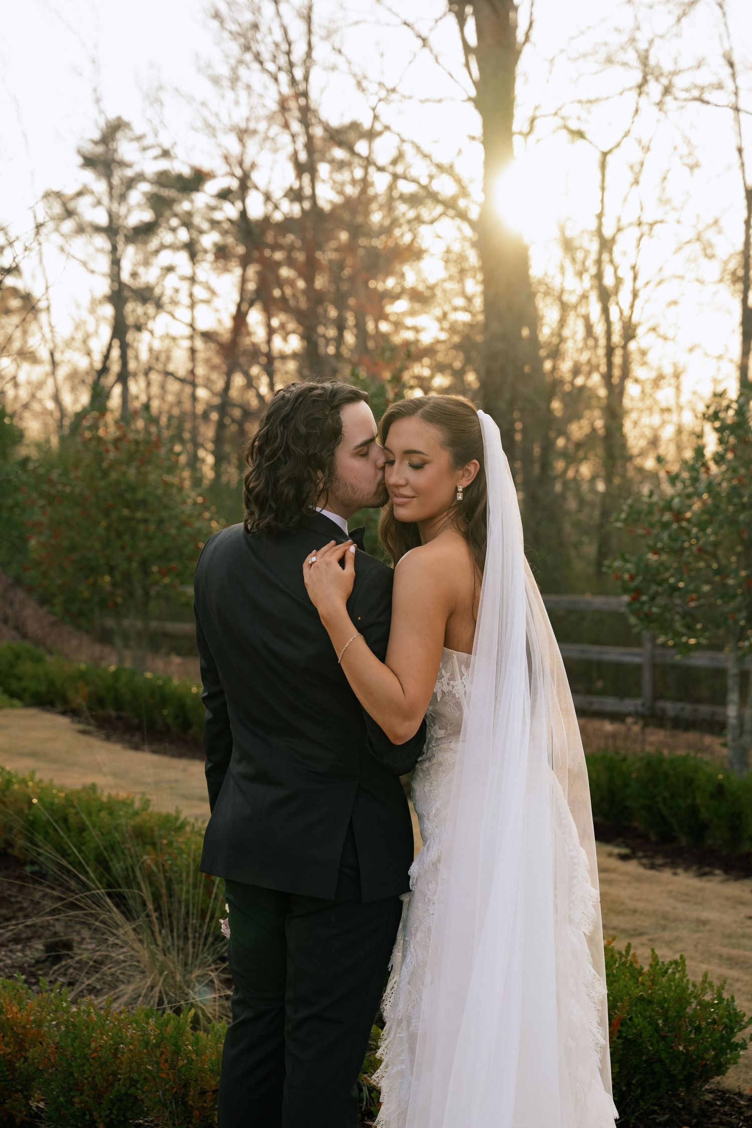 Bride and groom share a romantic kiss at golden hour in a garden, bride wearing a white gown and veil.