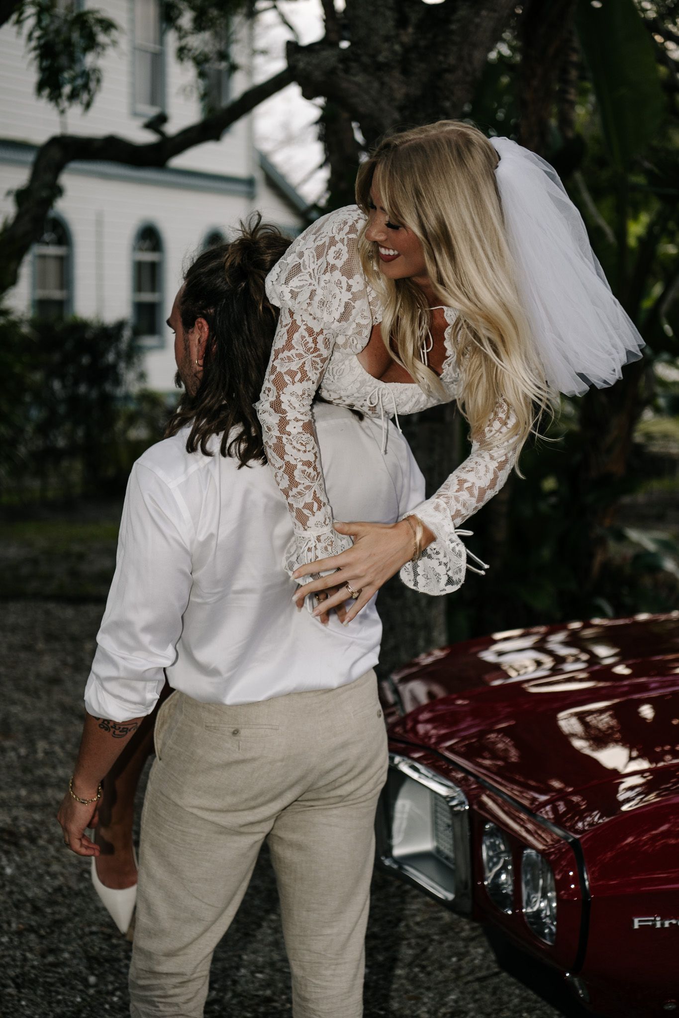 Couple posing playfully near vintage red car in casual wedding attire.