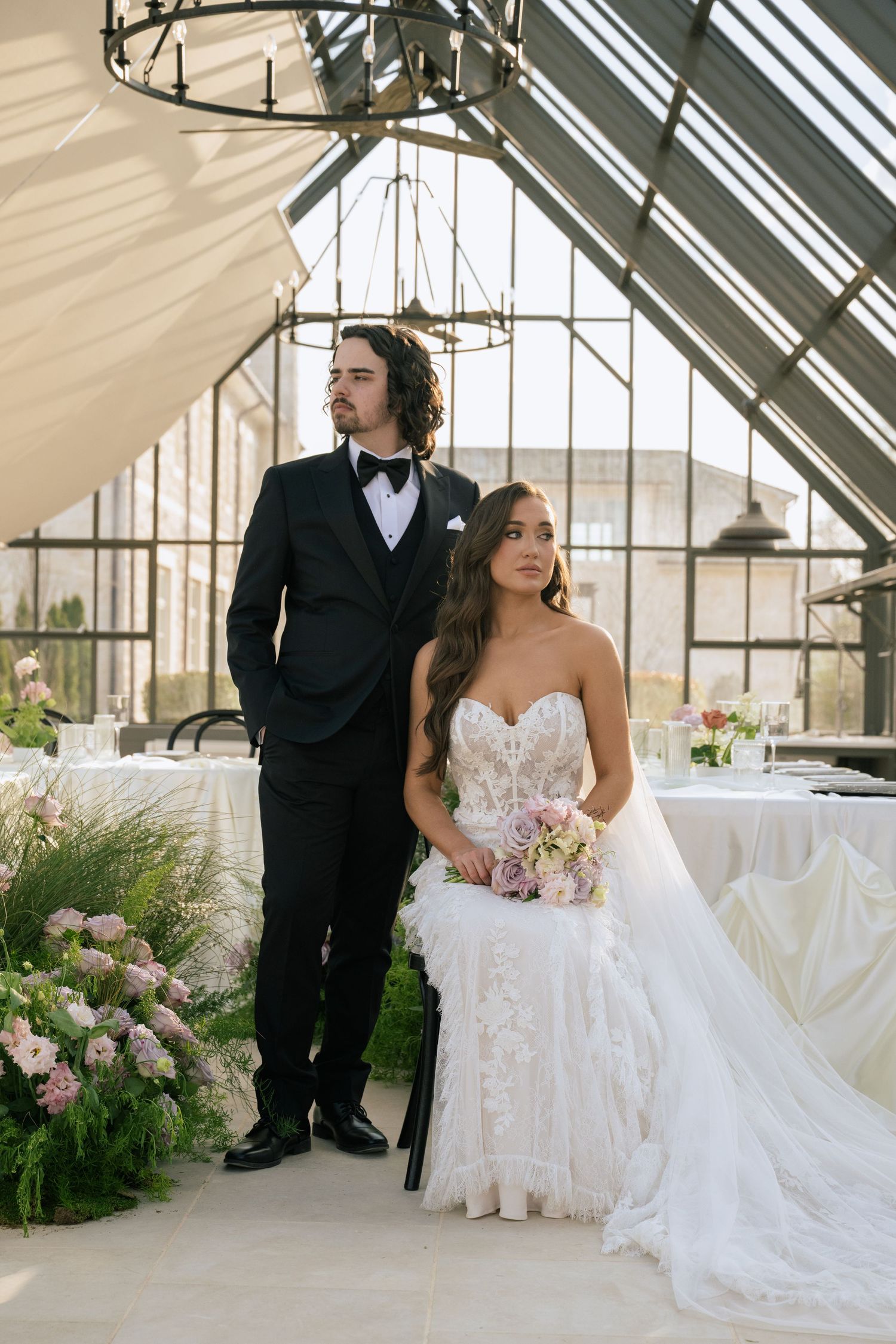 Bride in lace gown holding bouquet sits beside groom in black tuxedo inside elegant greenhouse wedding venue.