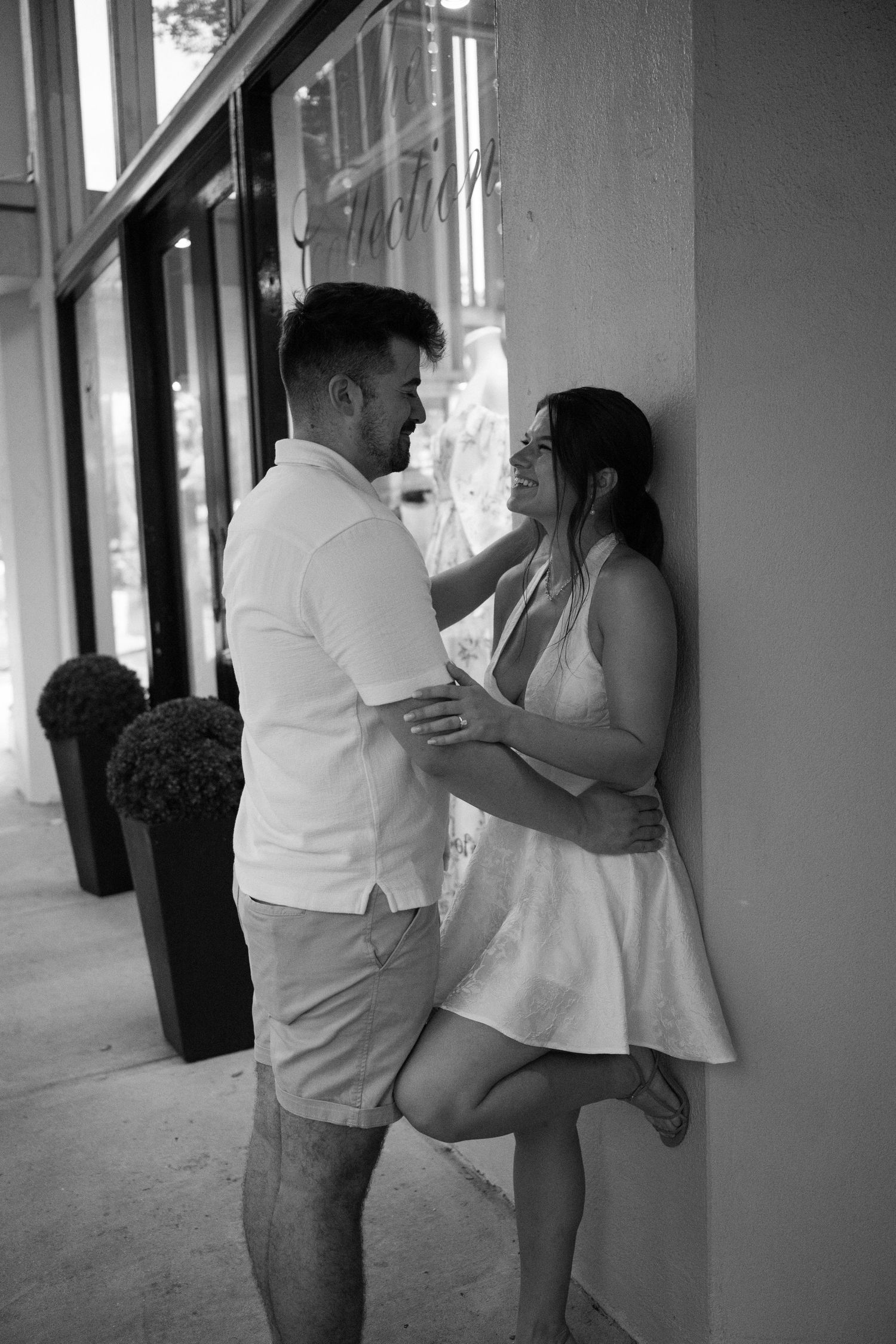Black and white photo of a romantic couple embracing against a wall outside a boutique shop.
