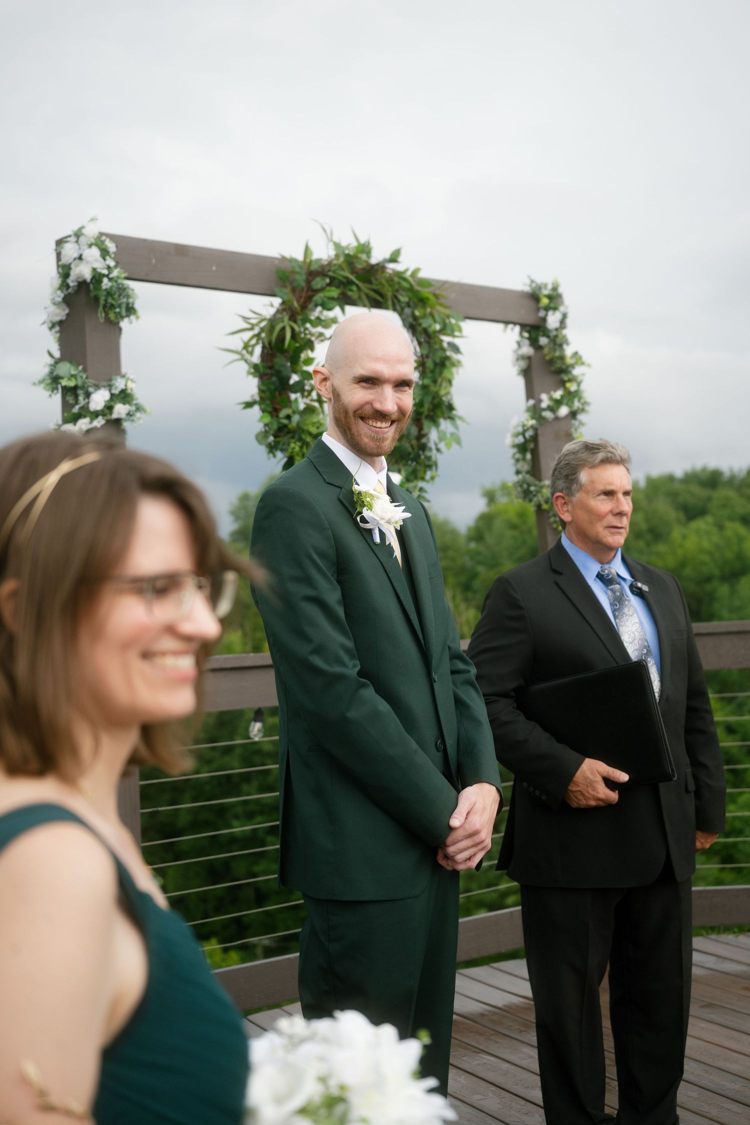 Groom in green suit smiling at altar during outdoor wedding ceremony with floral arch backdrop.