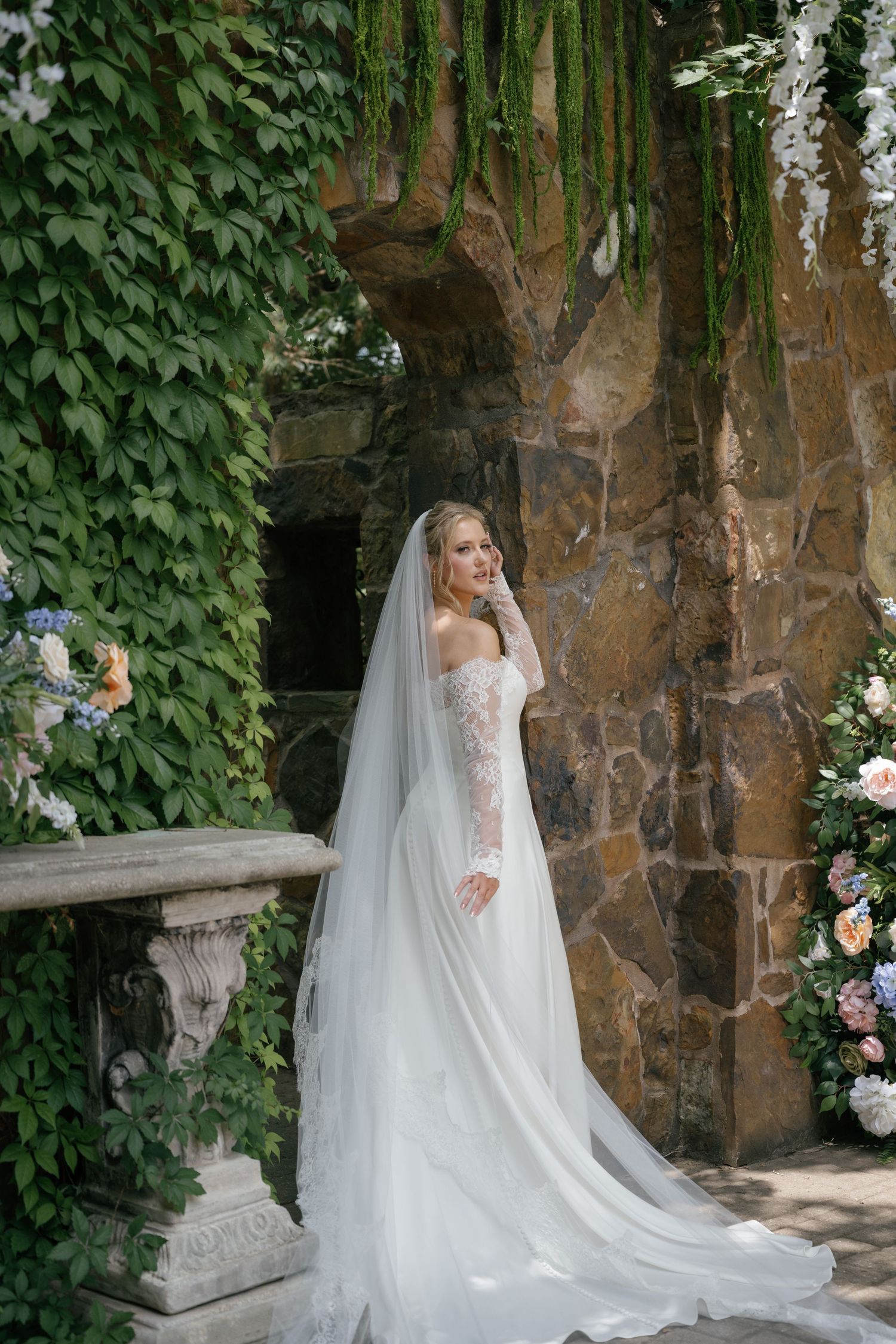 Elegant bride in lace off-shoulder gown and long veil poses by stone arch covered in lush ivy and floral arrangements.