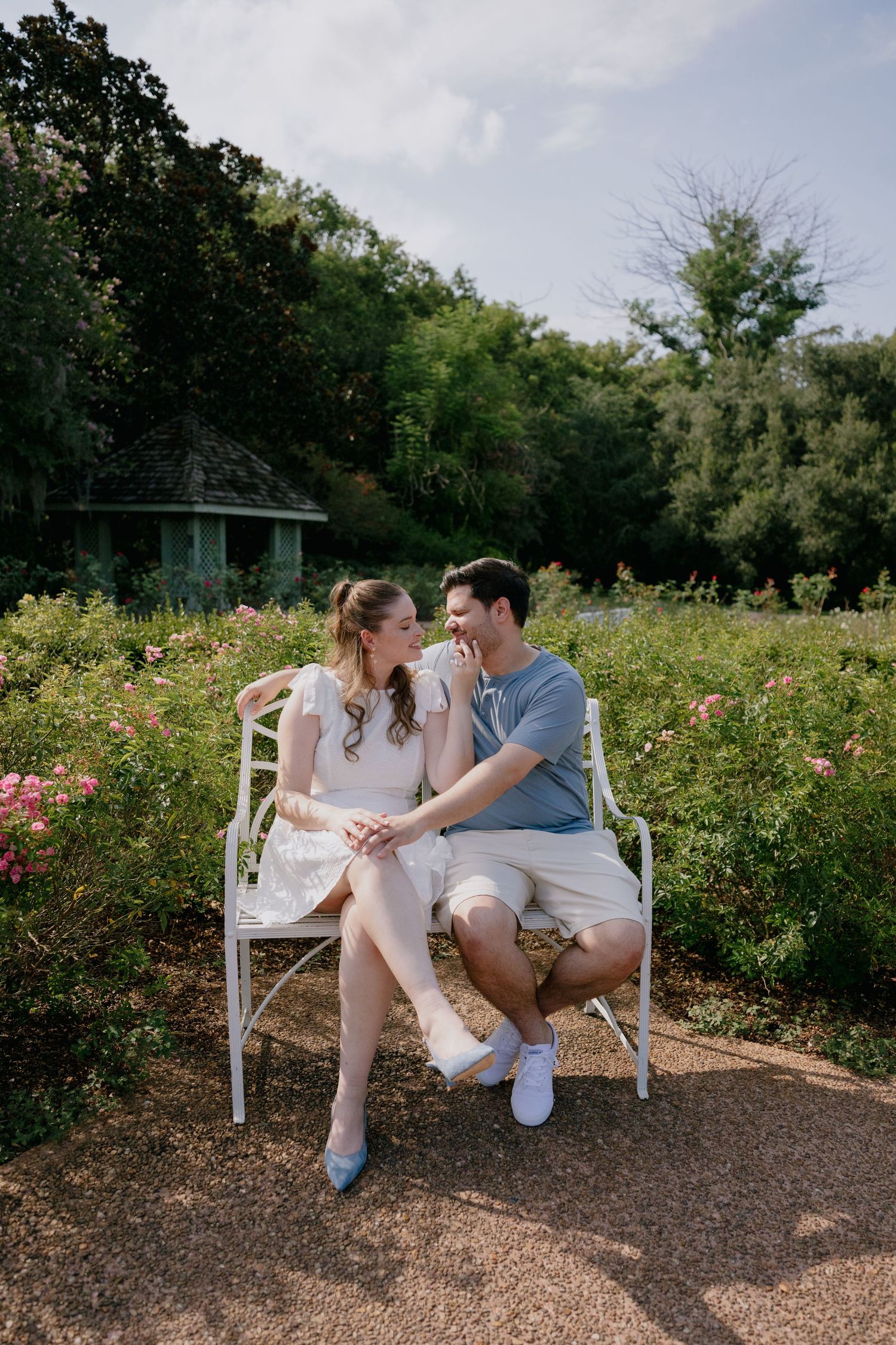 Couple sitting on white bench in rose garden, smiling at each other, surrounded by lush greenery and pink blooms.