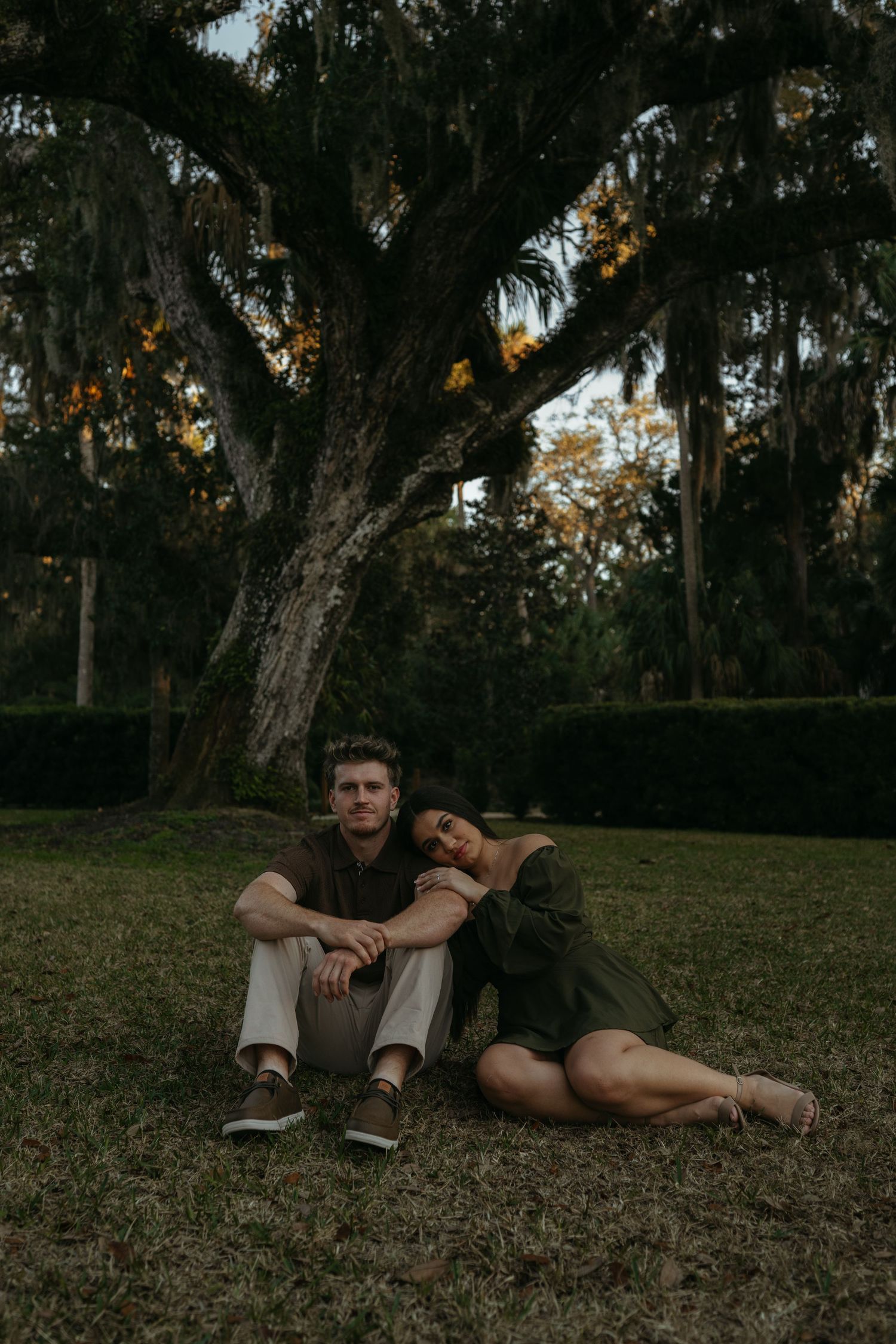 A person and their dog relax together on grassy ground under oak trees at dusk.
