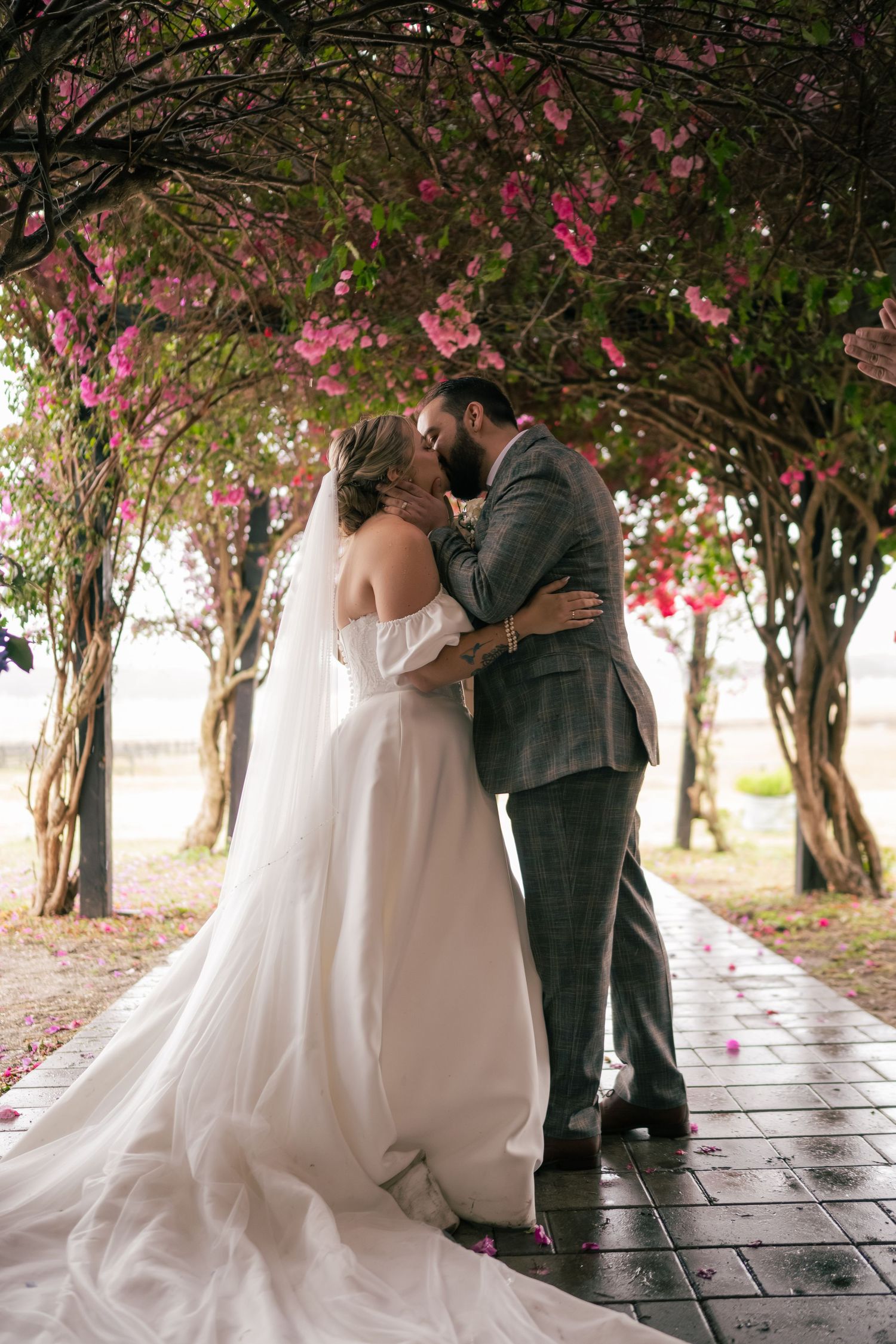A romantic wedding couple shares intimate moments under blooming cherry blossom trees in a garden walkway.