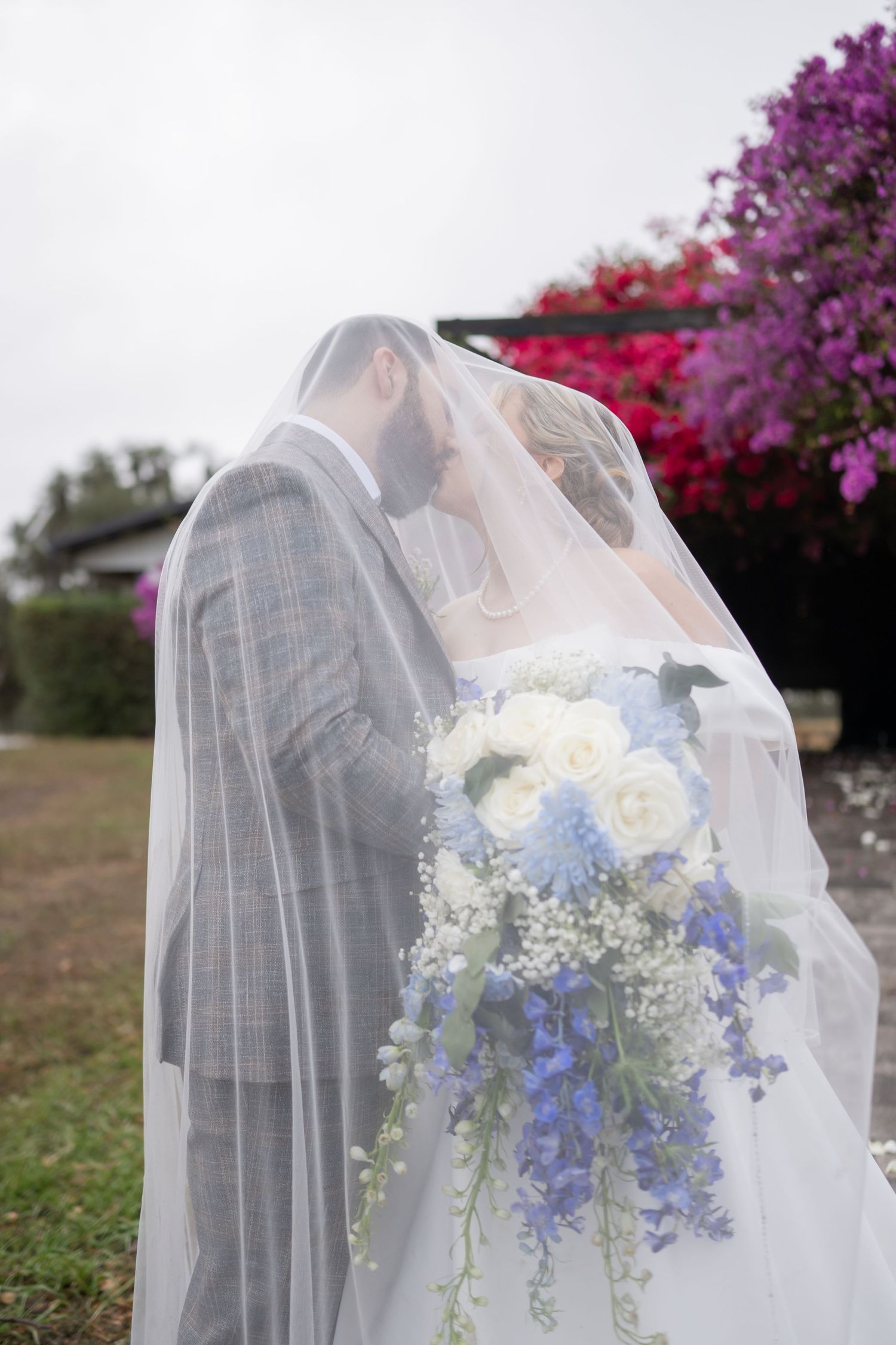 Close-up of a delicate white and blue wedding bouquet with roses and baby's breath behind a flowing veil.