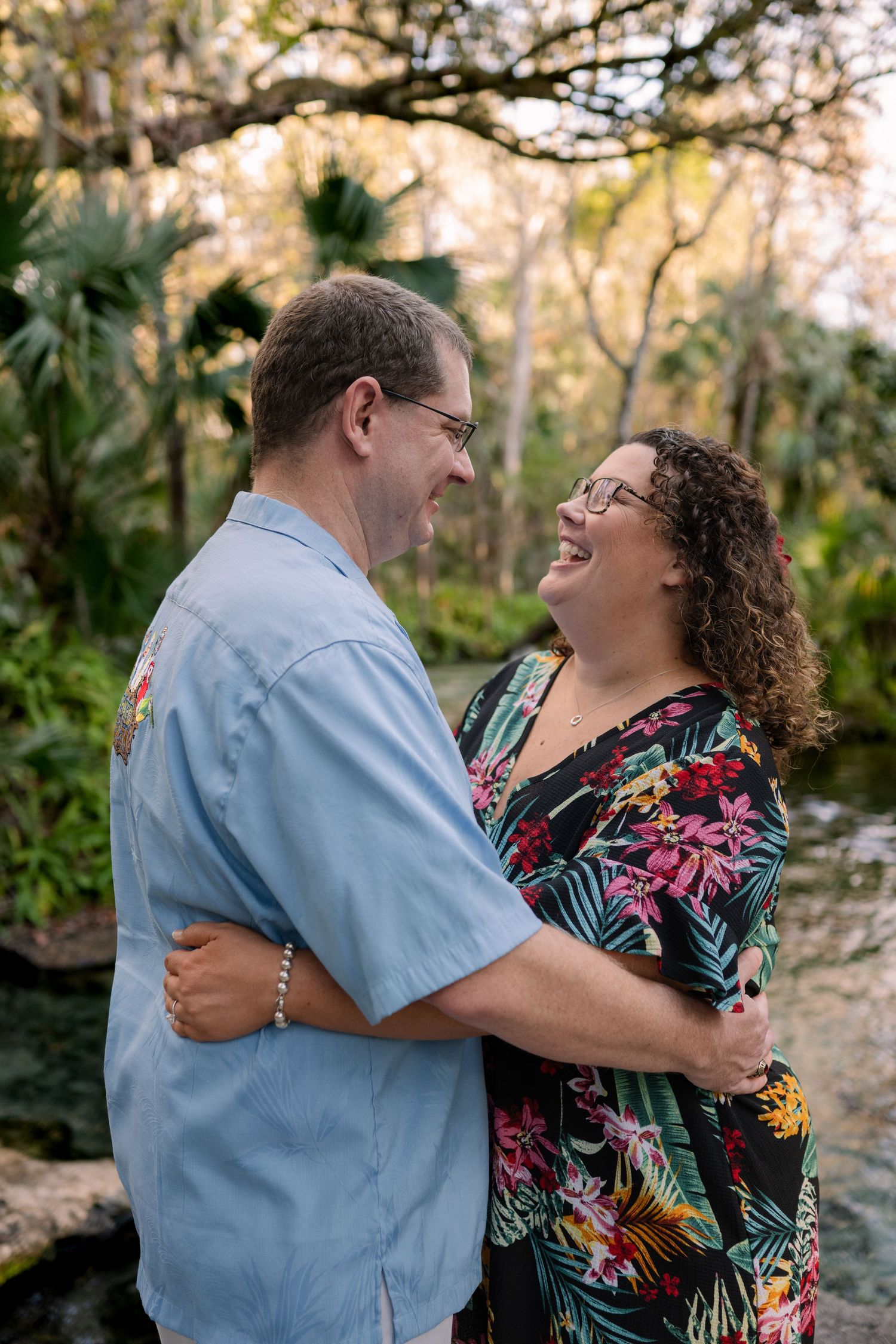 A couple in tropical attire laughs and embraces outdoors in a lush garden setting during golden hour.