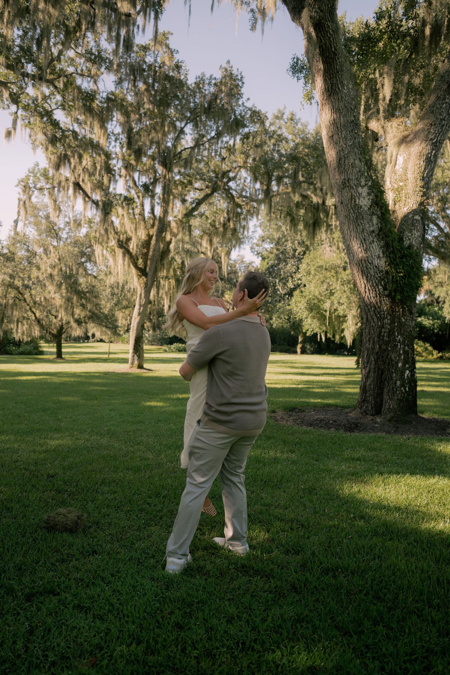 Couple embracing and dancing outdoors under Spanish moss-draped oak trees on lush green lawn at golden hour.