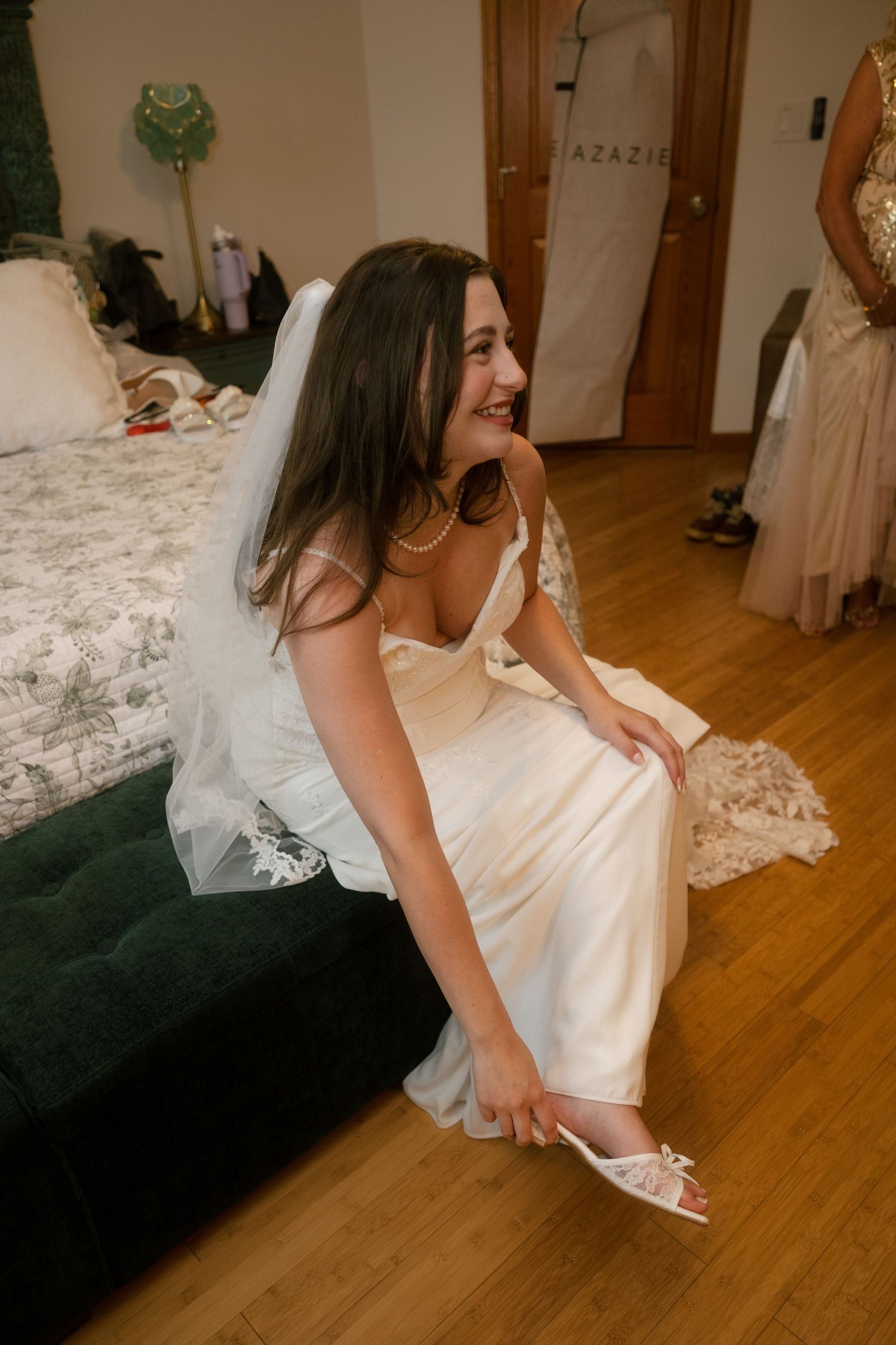 Bride in white gown and veil sits on green ottoman putting on heels, smiling joyfully while getting ready.