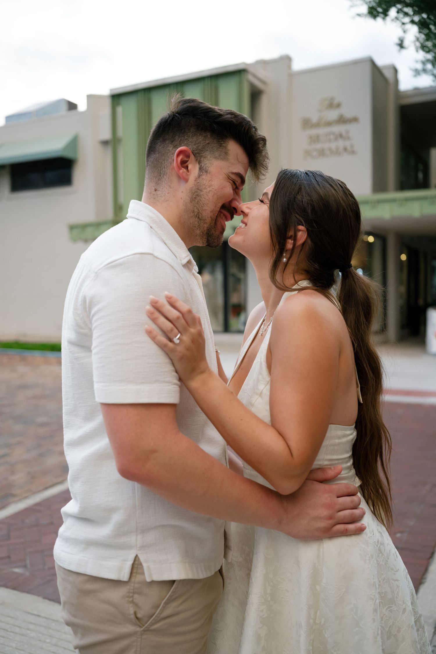 A couple in white outfits shares a romantic moment outside The Ballerina Bridal Formal shop.
