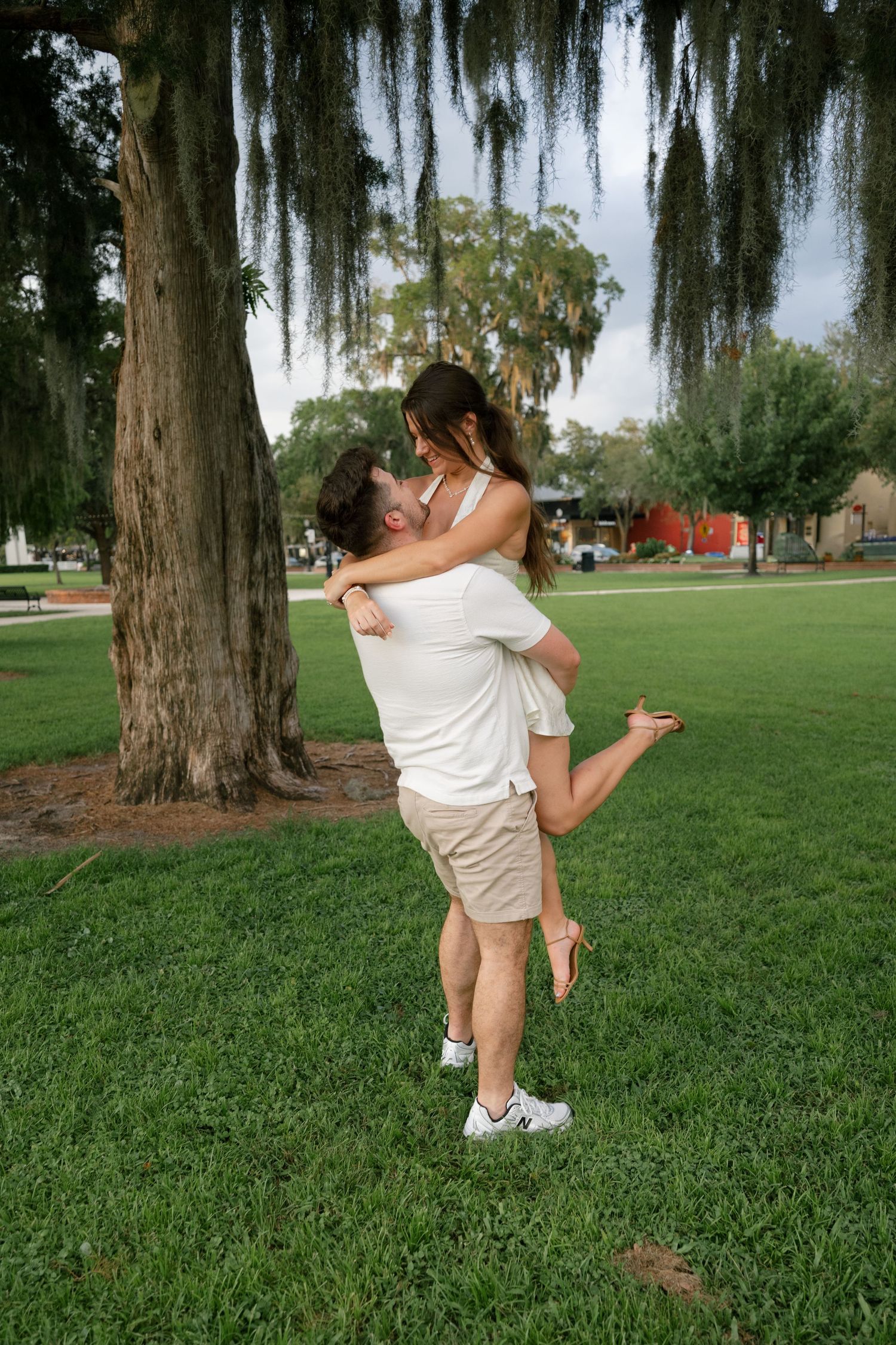 A man lifts and embraces a woman near a large tree in a lush green park, sharing a romantic moment.