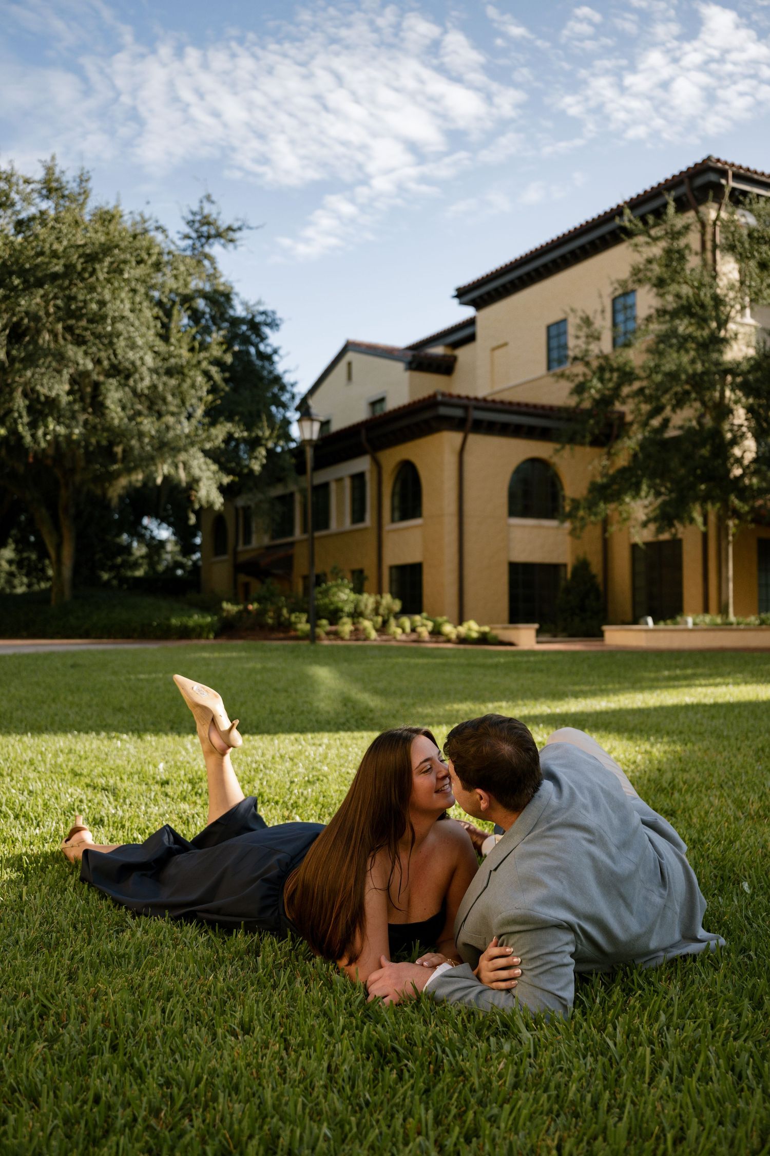 A couple lies close together on a sunny green lawn, sharing an intimate moment in front of a tan Mediterranean-style building.