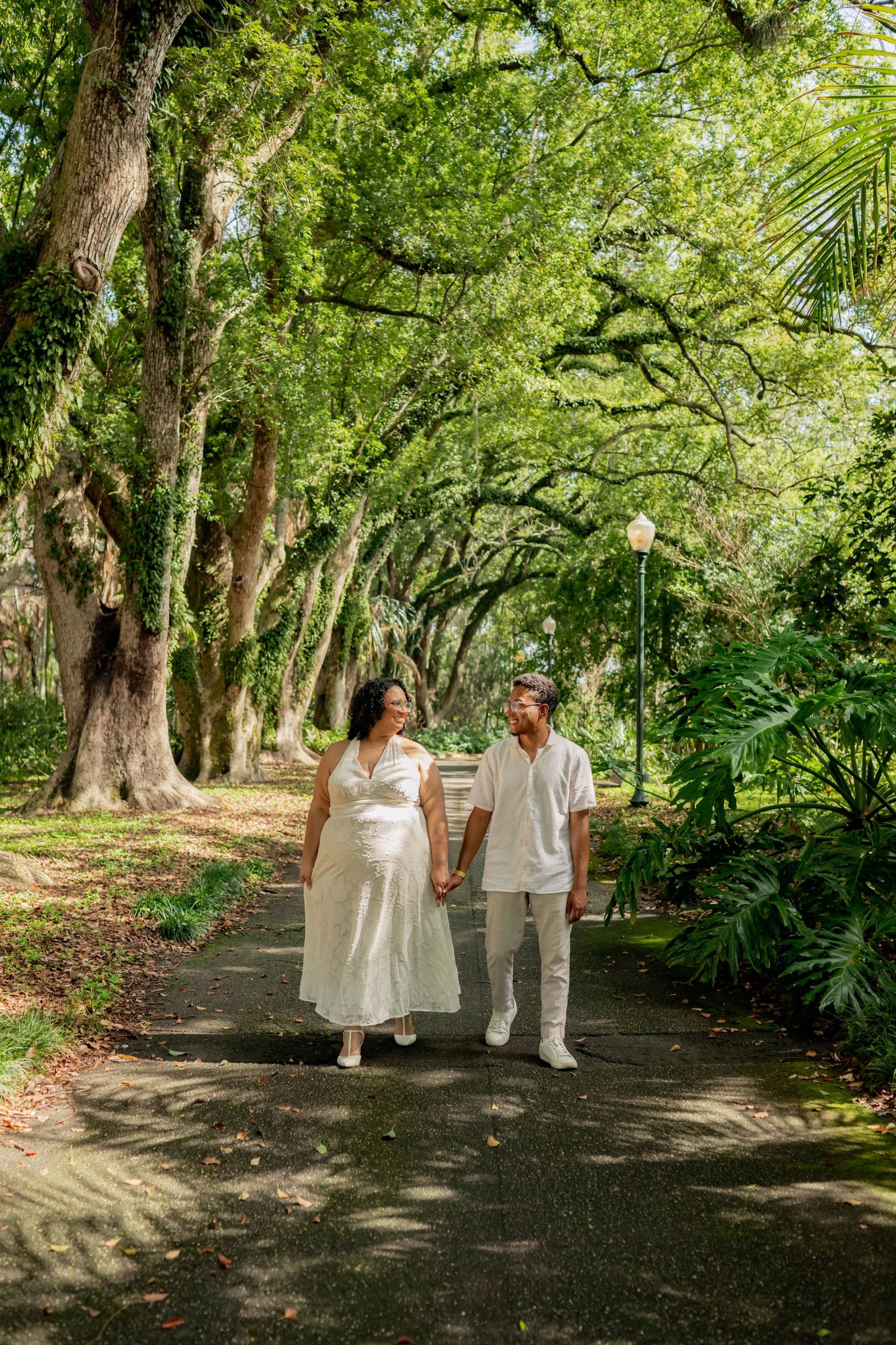Couple in white attire walking hand-in-hand on a shaded park path lined with lush green trees.