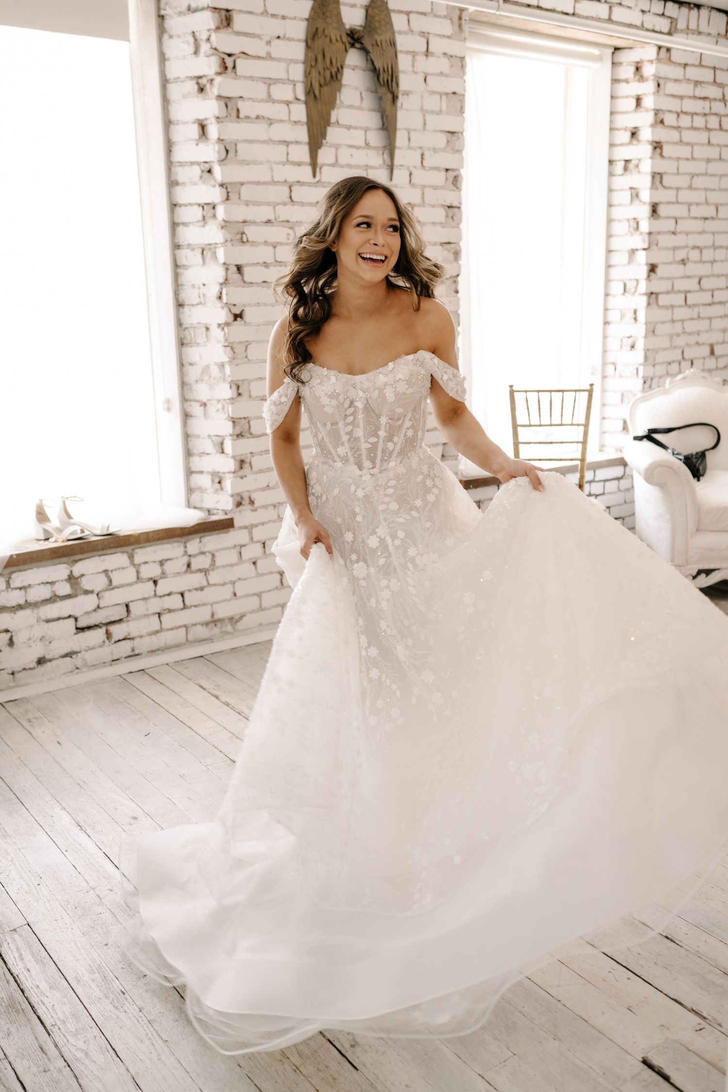 Smiling bride in off-shoulder beaded ballgown twirling in bright white brick room with wood floors.