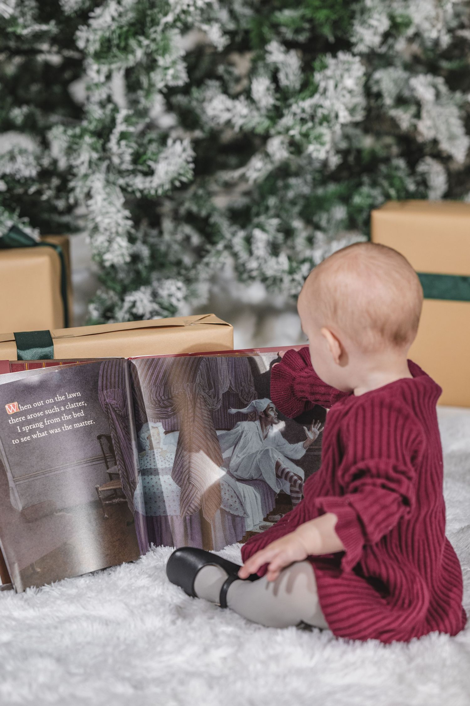 Baby in red sweater dress lies on snowy white setting with holiday decorations.