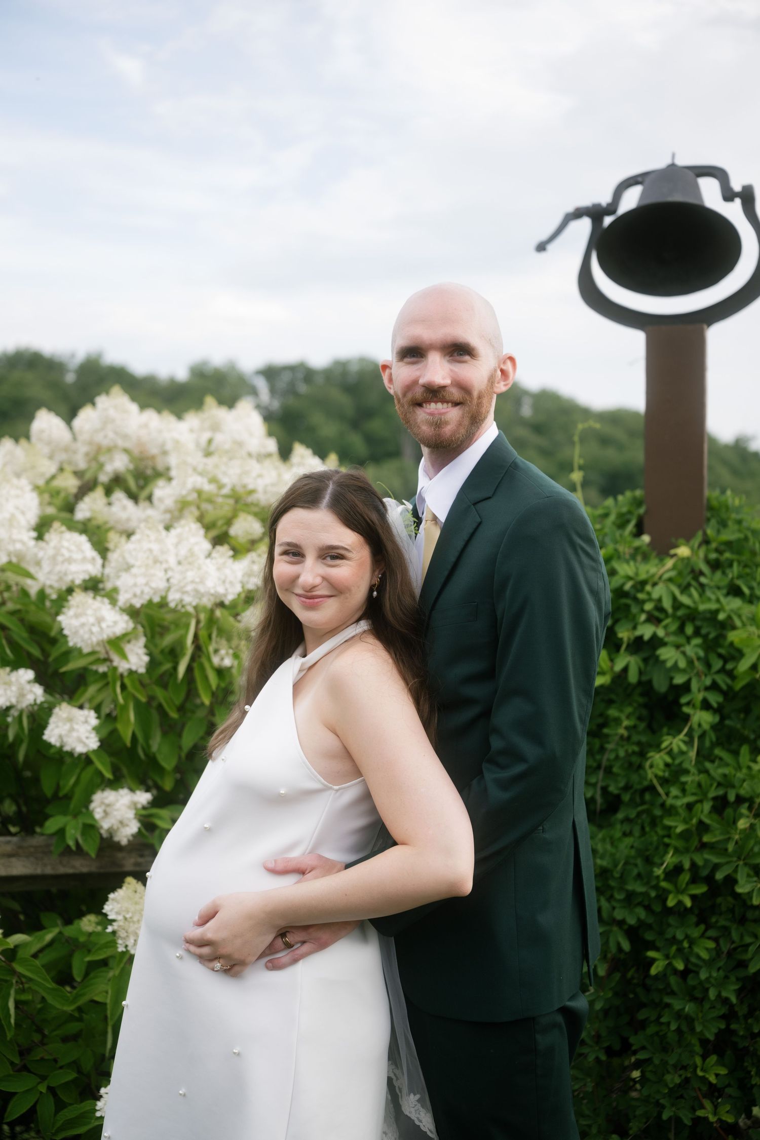 Pregnant bride in white dress embraces groom in green suit outdoors with white hydrangeas blooming behind them.