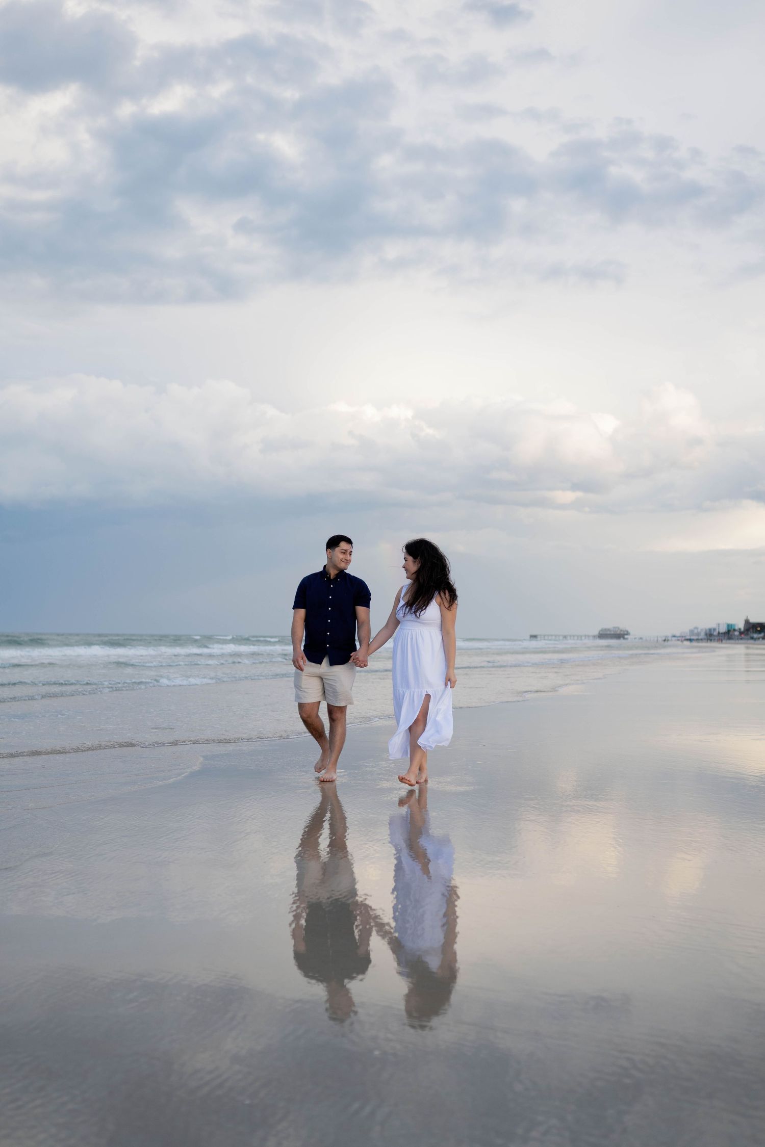 Couple holding hands walking on a reflective wet beach at sunset with dramatic cloudy sky.