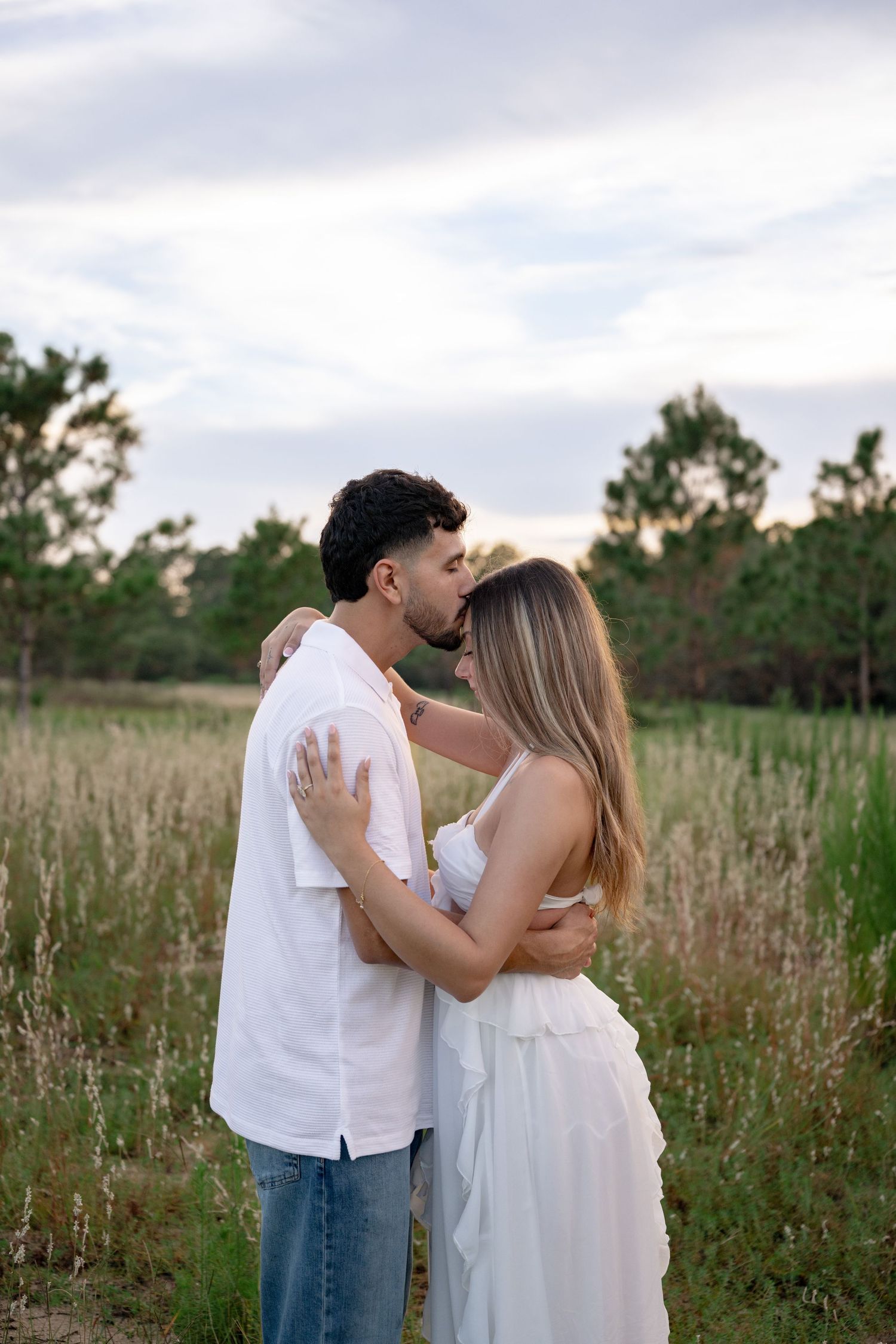 Couple embracing romantically in a field at sunset, woman in white dress, man in white shirt and jeans.