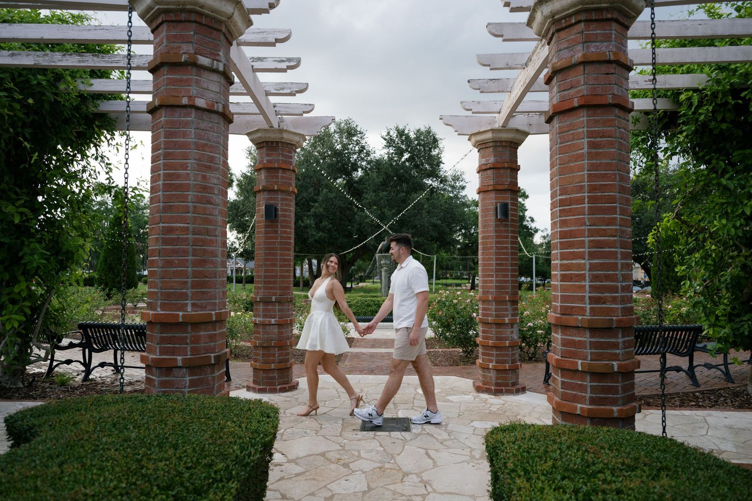 Couple holding hands walking through brick pergola columns in a lush garden park setting.